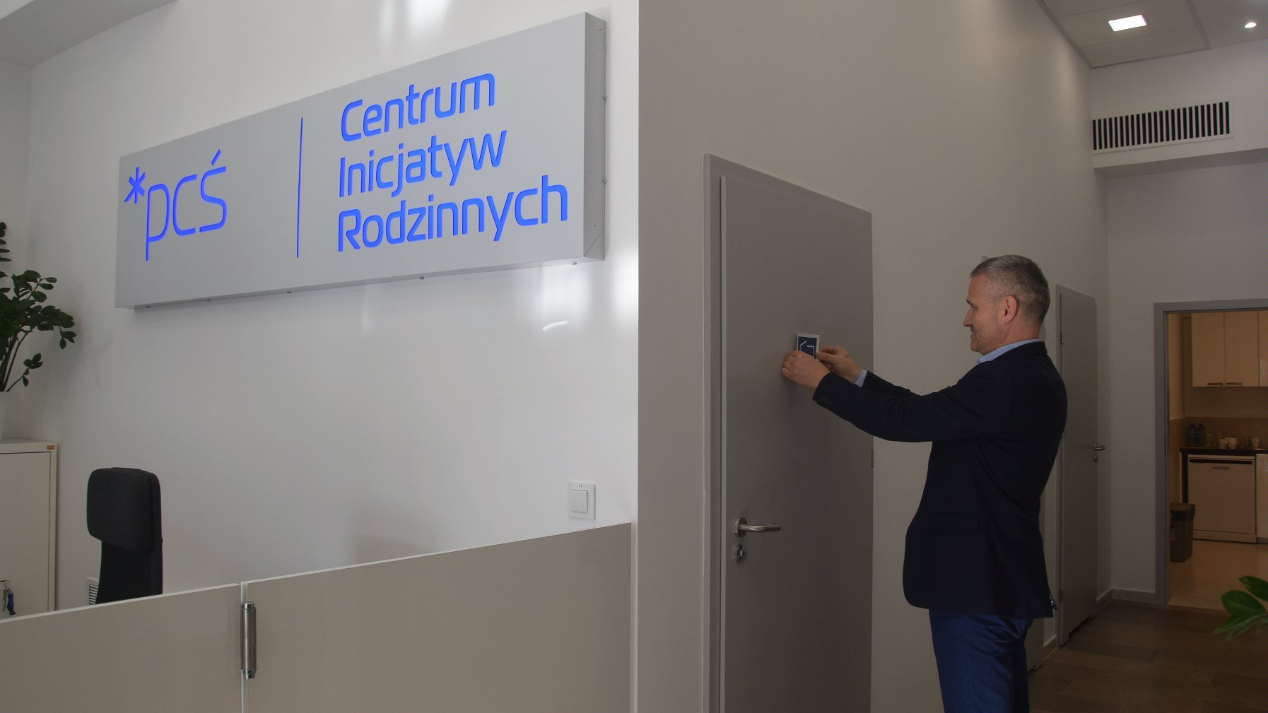 Wnętrze wyremontowanej siedziby Centrum Inicjatyw Rodzinnych. Na białej ścianie niebieskie logo centrum. Na drzwiach wiceprezydent Poznania Jędrzej Solarski zakłada oznaczenie miejsca, w którym można przewinąć dorosłą osobę pieluchowaną.