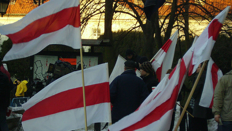 Grupa ludzi odwróconych tyłem, trzymają białoruskie flagi (biało-czerwono-białe); na pierwszym planie kolejne białoruskie flagi - grafika artykułu