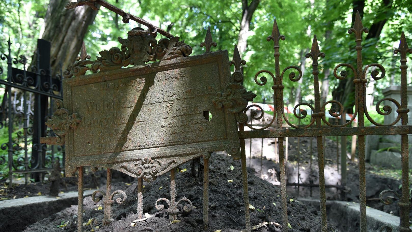 Metalowy, kuty nagrobek. Na pierwszym planie ozdobna krata i metalowa tablica nagrobna, w tle drzewa.