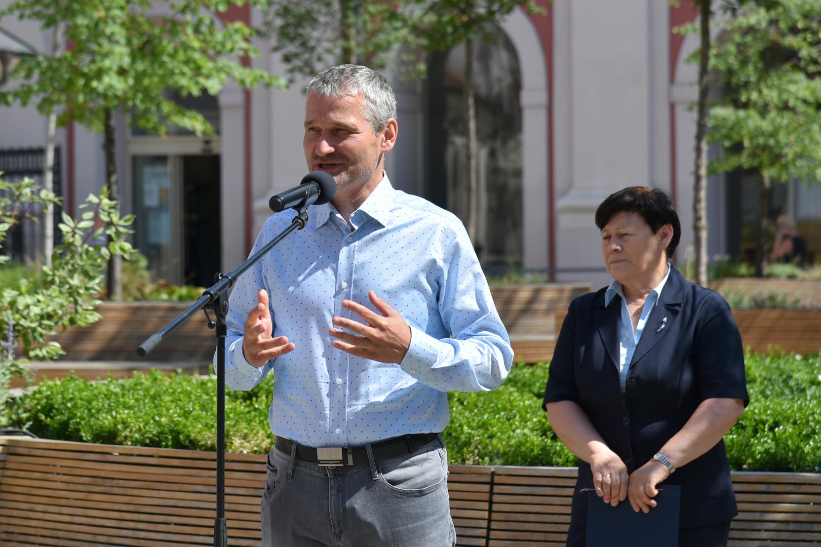 Jędrzej Solarski, zastępca prezydenta Poznania, stoi przy mikrofonie na dziedzińcu urzędu. Za nim Ewa Bąk, dyrektor Wydziału Sportu UMP. W tle urząd miasta i rośliny - grafika artykułu