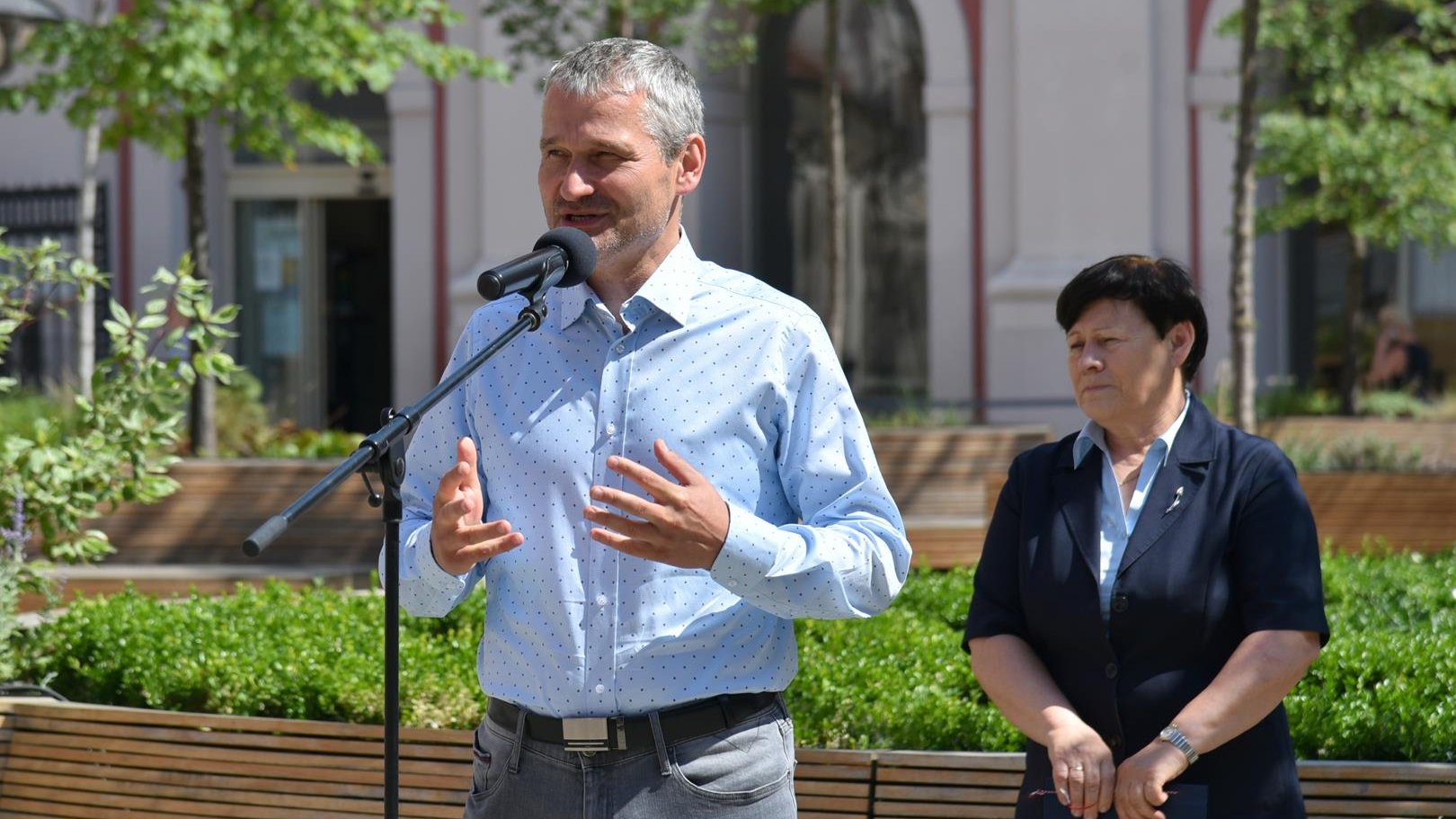 Jędrzej Solarski, zastępca prezydenta Poznania, stoi przy mikrofonie na dziedzińcu urzędu. Za nim Ewa Bąk, dyrektor Wydziału Sportu UMP. W tle urząd miasta i rośliny