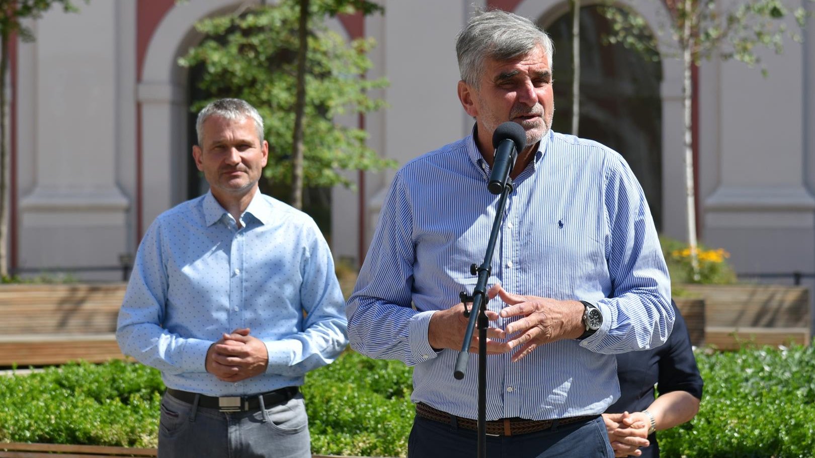 Ryszard Stadniuk, prezes Polskiego Związku Towarzystw Wioślarskich stoi przy mikrofonie na dziedzińcu urzędu. Za nim stoi Jędrzej Solarski, zastępca prezydenta Poznania. W tle budynek urzędu i drzewa