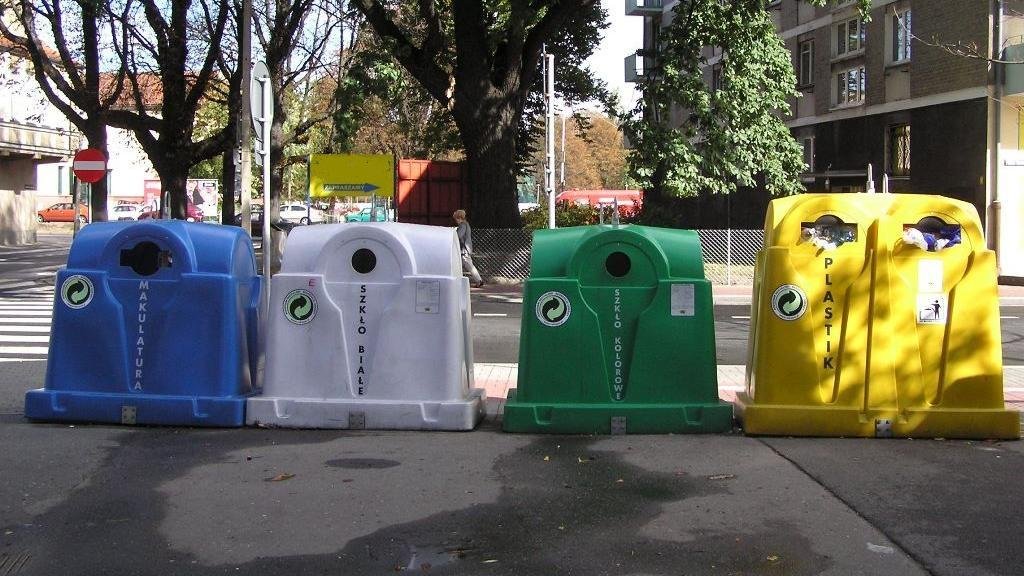 Na zdjęciu znajdują się cztery kolorowe pojemniki na odpady (niebieski, biały, zielony i żółty) stojące na chodniku, przy ulicy.