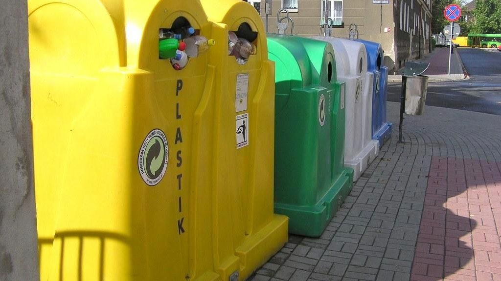 Na zdjęciu znajdują się cztery kolorowe pojemniki na odpady (żółty, zielony, biały i niebieski) stojące na chodniku, przy ulicy.