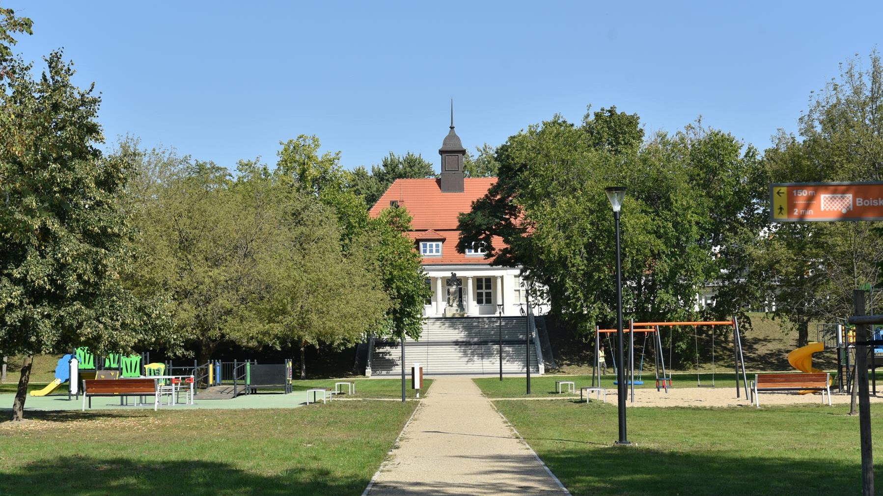 Zdjęcie przedstawia jeden z budynków Dziecińca pod Słońcem. Obiekt znajduje się za drzewami. Na pierwszym planie widać place zabaw.