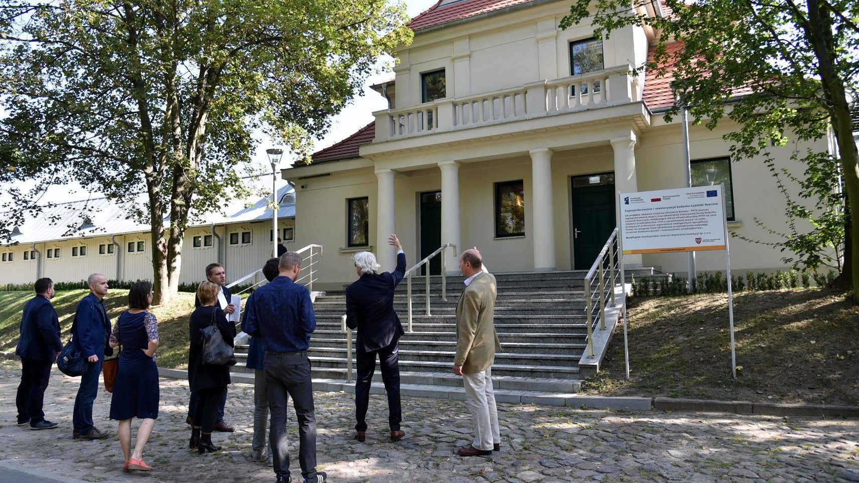 Zdjęcie przedstawia budynek Łazienek Rzecznych z zewnątrz. Przed budynkiem stoją przedstawiciele Miasta Poznania i zarządcy budynku.