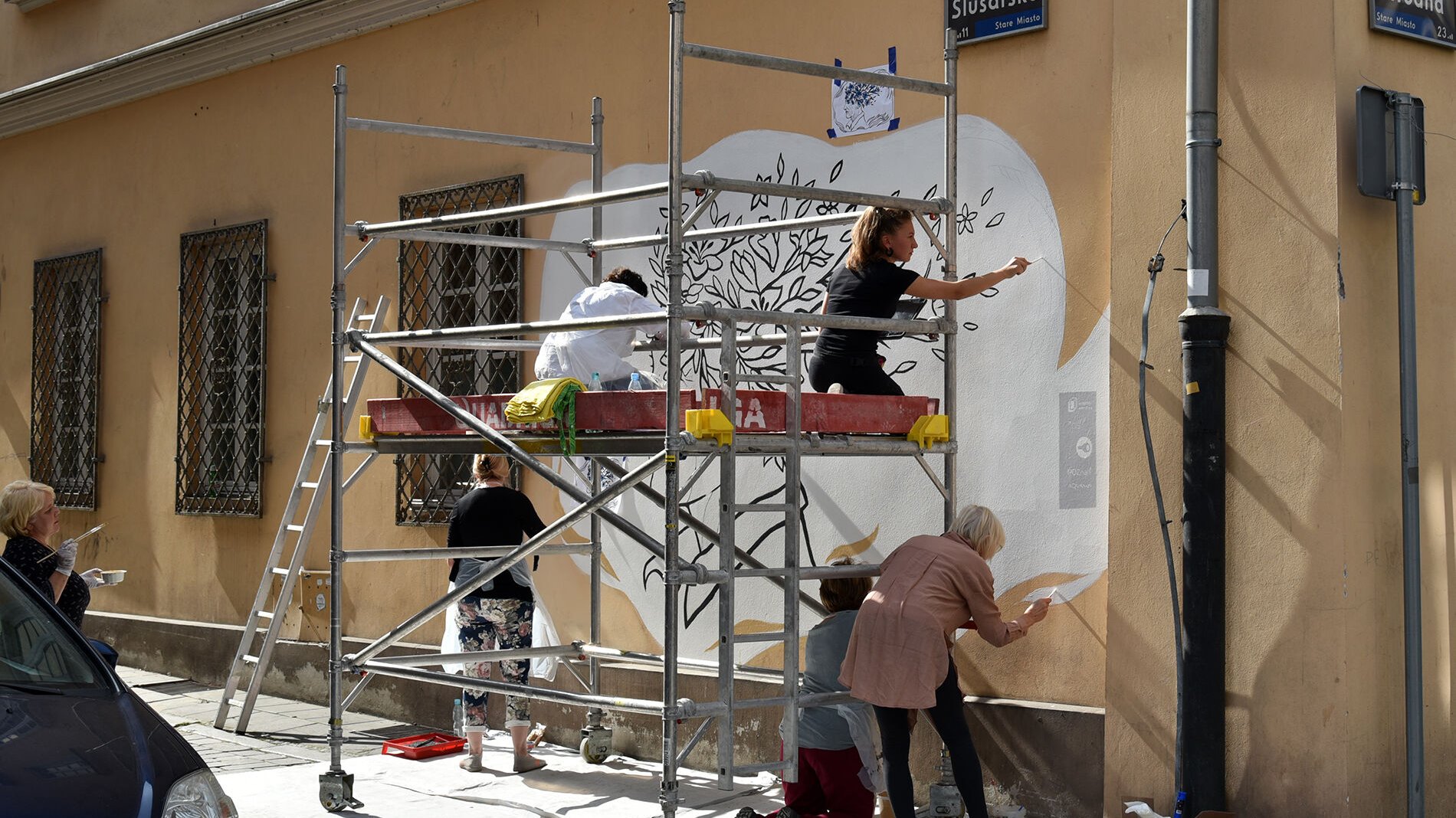 Widok na róg ulicy. Pod ścianą budynku rusztowanie, na nim pięć kobiet maluje mural. Szósta stoi za nimi, przygląda się pracy z odległości kilku kroków