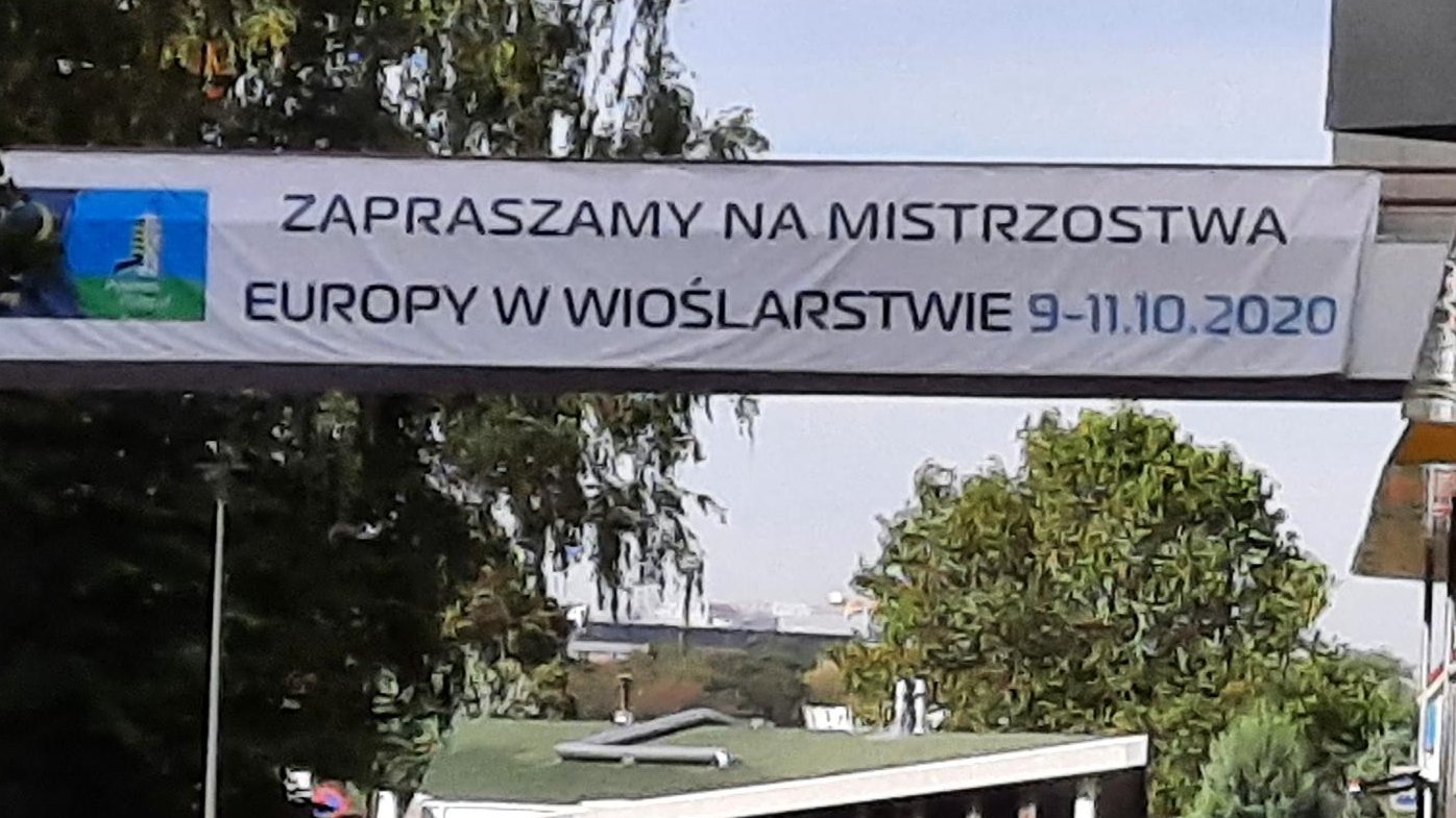 Kadr z widokiem na tor regatowy. Między drzewem a budynkiem rozpięty baner z napisem: Zapraszamy na Mistrzostwa Europy w Wioślarstwie, 9-11.10.2020