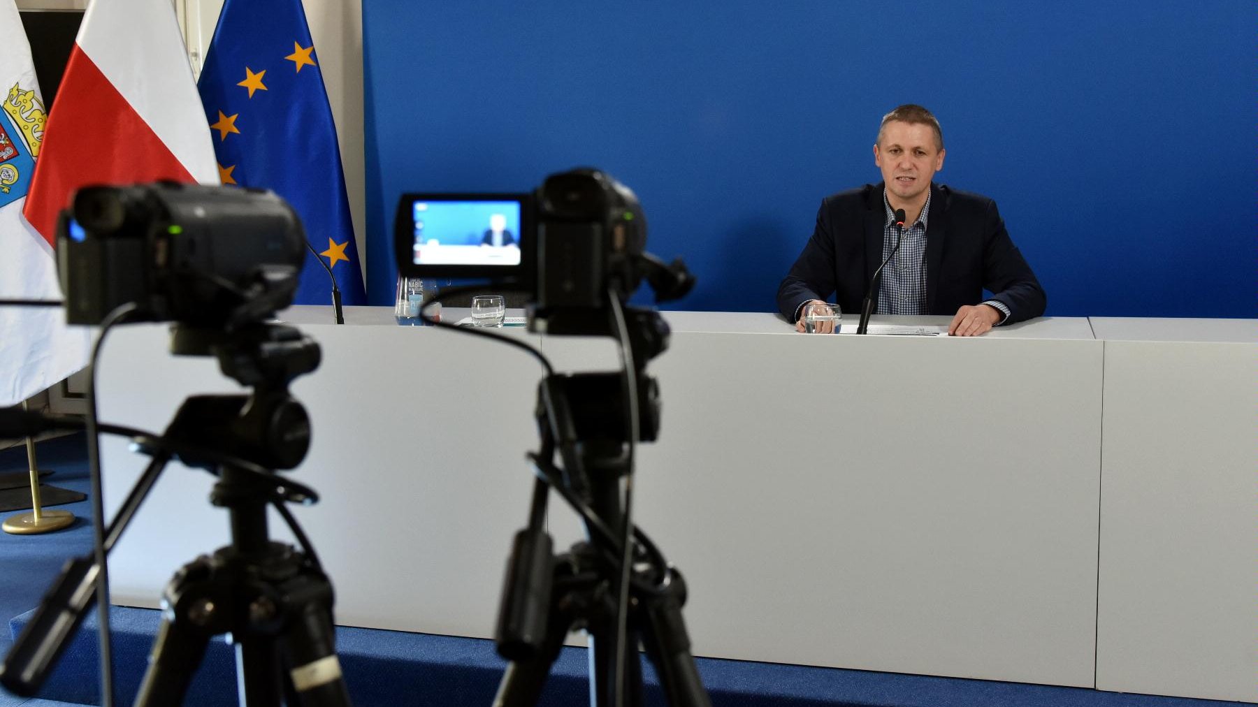 Zdjęcie przedstawia Patryka Pawełczaka, dyrektora Gabinetu Prezydenta Urzędu Miasta za stołem konferencyjnym. Na pierwszym planie widać kamery.