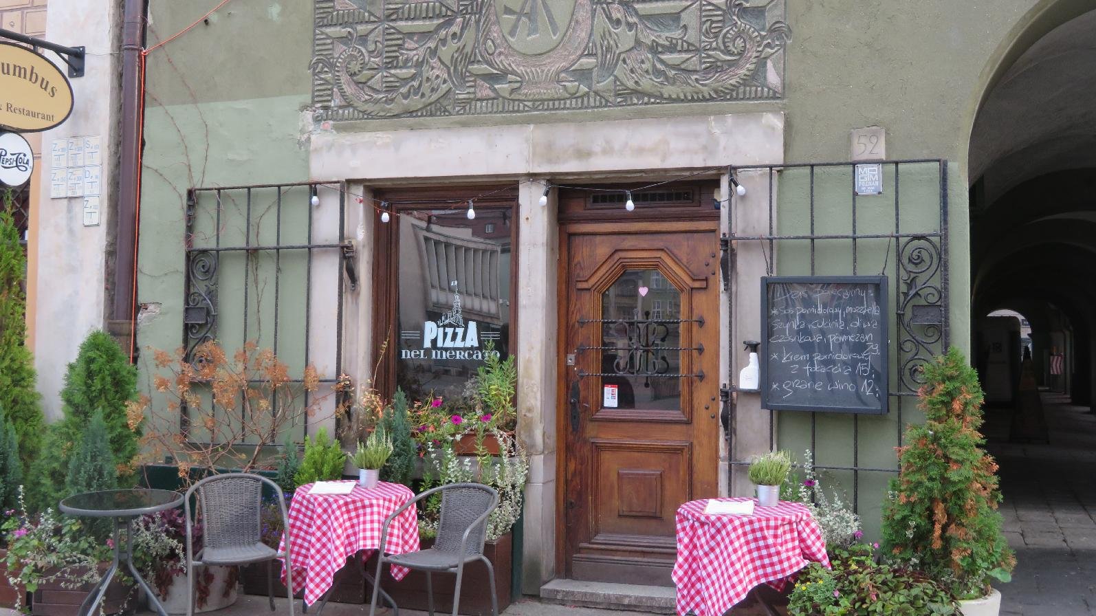 Zdjęcie przedstawia budynek pizzerii. Przed lokalem stoją stoliczki z czerwono - białym obrusem w kratkę i krzesła.