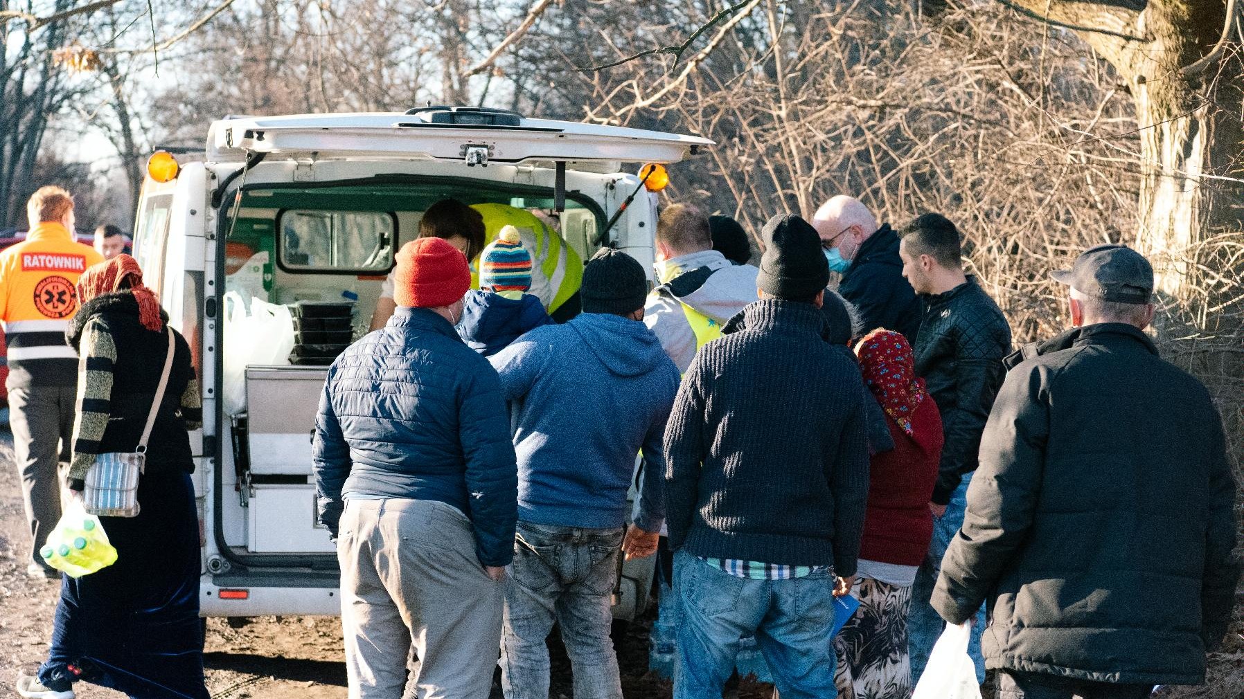 Ludzie stoją w kolejce przed samochodem dostawczym, z którego wydawane są im posiłki.