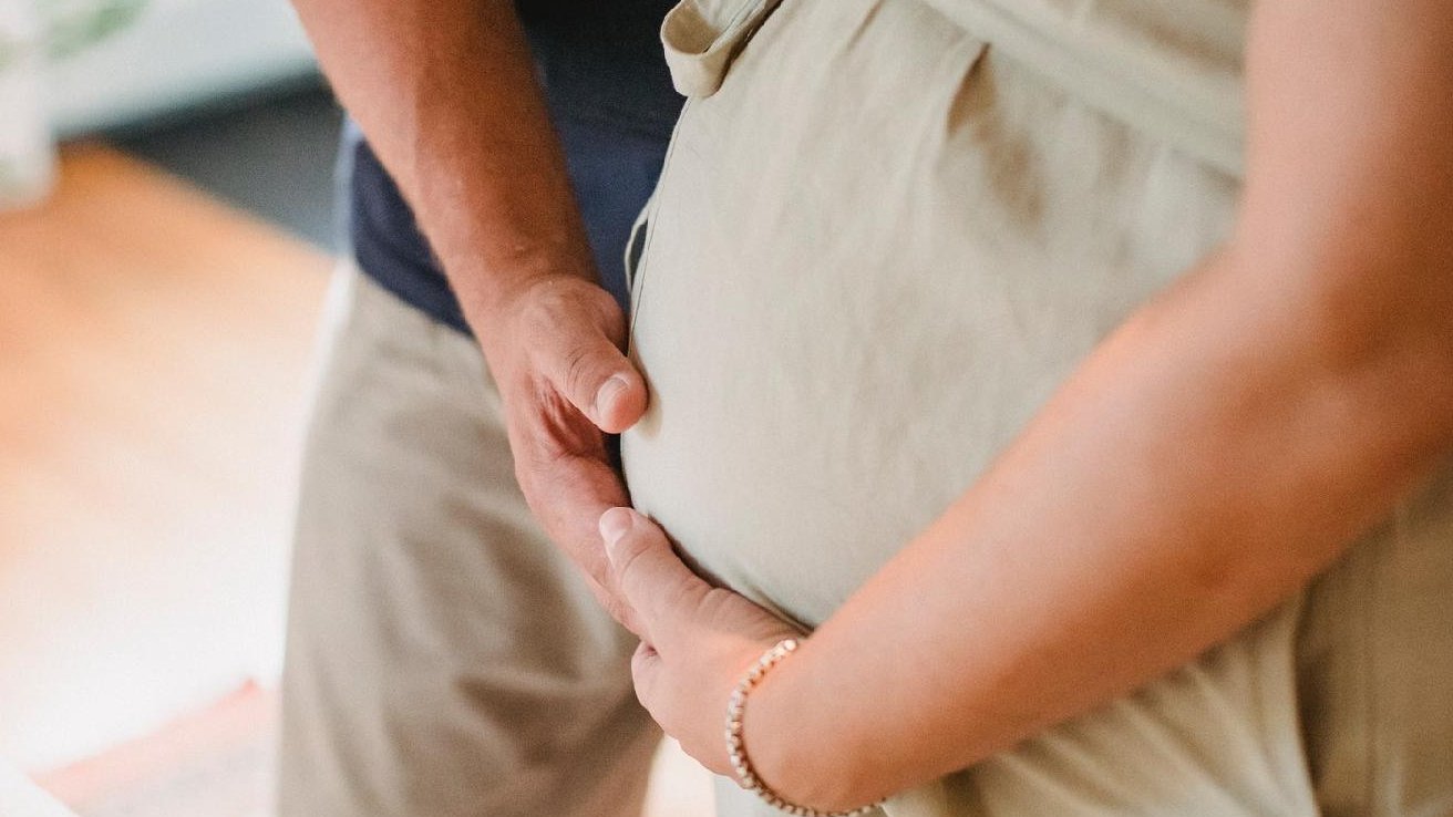Na zdjęciu mężczyzna i kobieta w ciąży, stoją obok siebie, trzymając dłonie na brzuchu kobiety