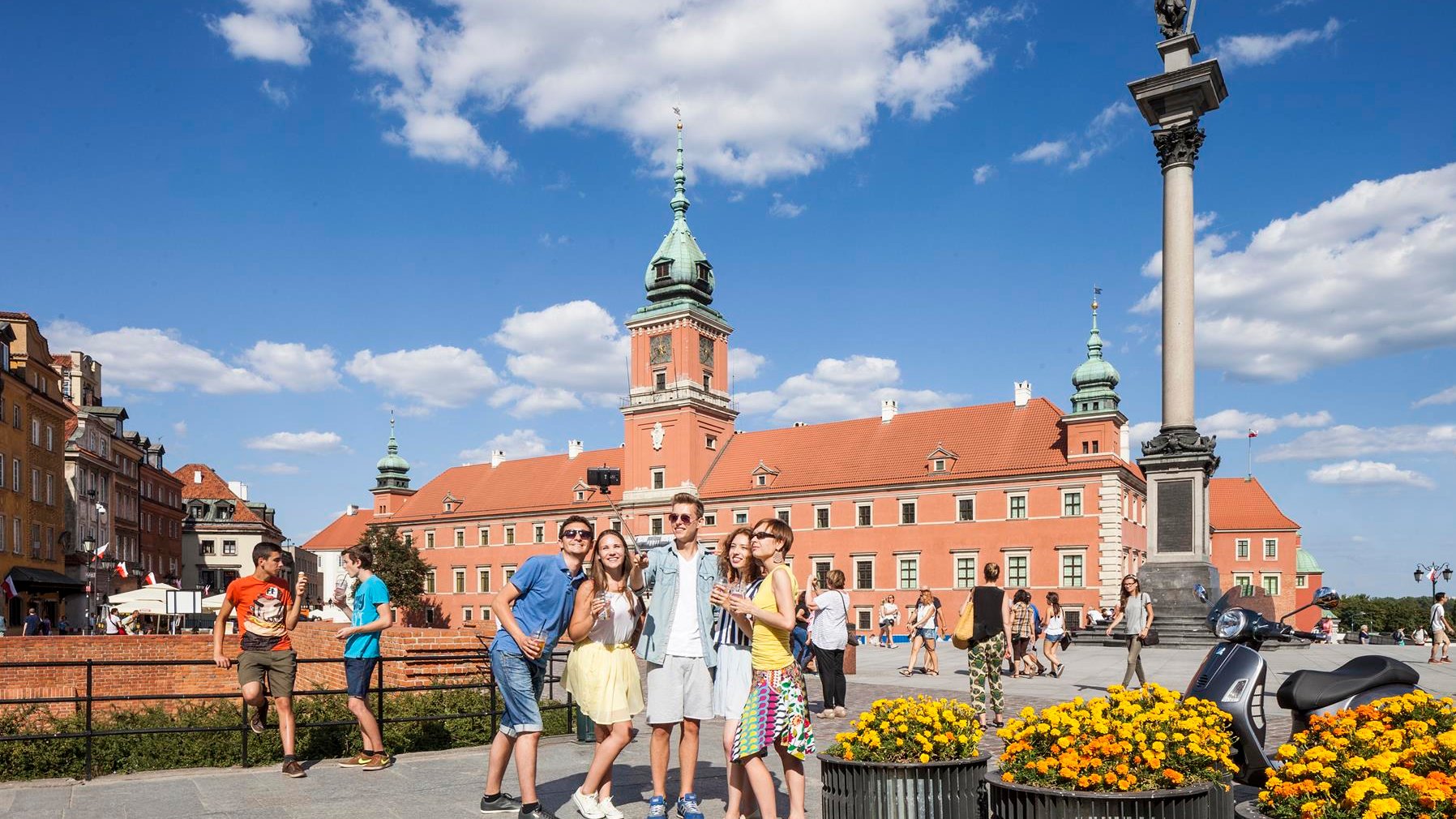Na zdjęciu plac Zamkowy w Warszawie, na pierwszym planie uśmiechnięci turyści