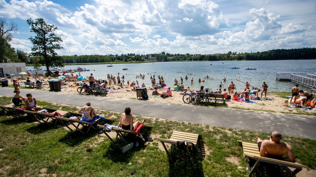 Zdjęcie przedstawia ludzi siedzących i leżących na plaży nad jeziorem.