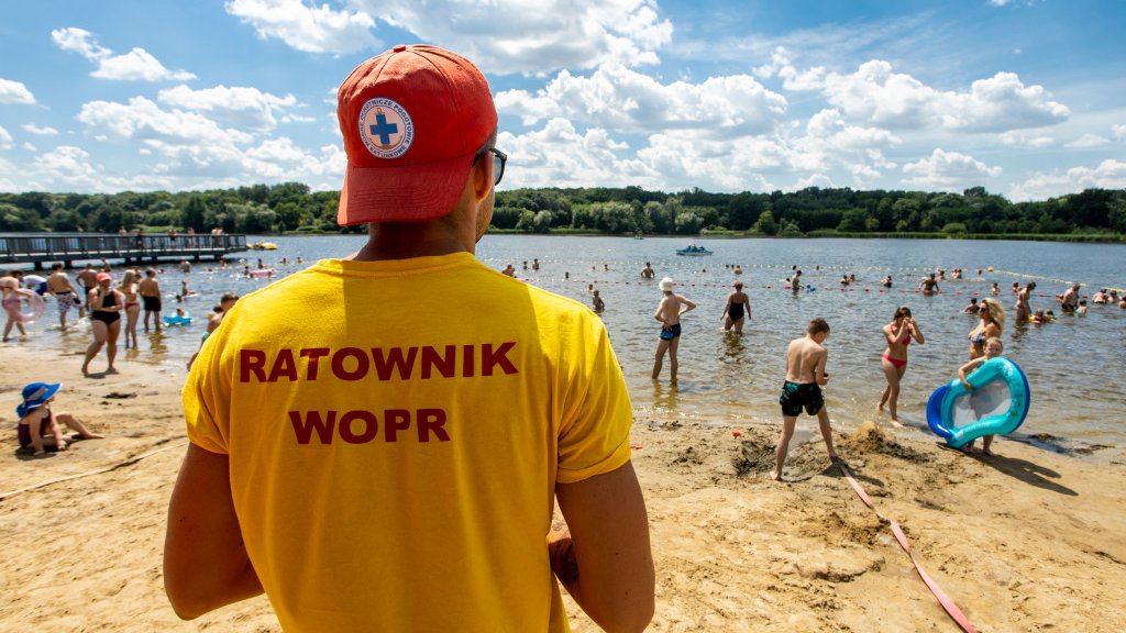 Zdjęcie przedstawia ludzi kąpiących się w jeziorze. Na pierwszym planie widać ratownika stojącego tyłem do aparatu i przodem do plaży.