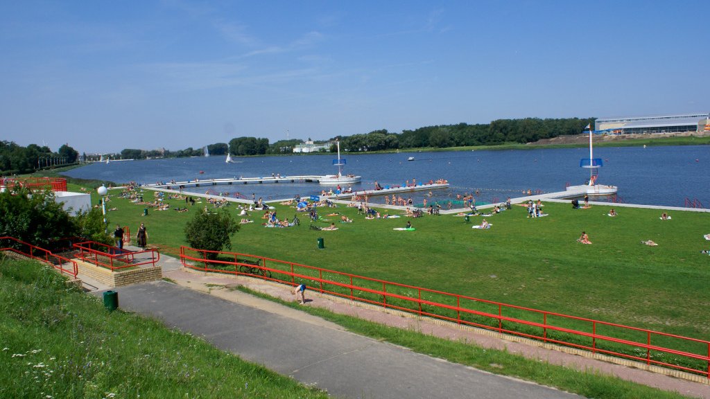 Zdjęcie przedstawia jezioro i trawiastą plażę, na której wypoczywają ludzie.
