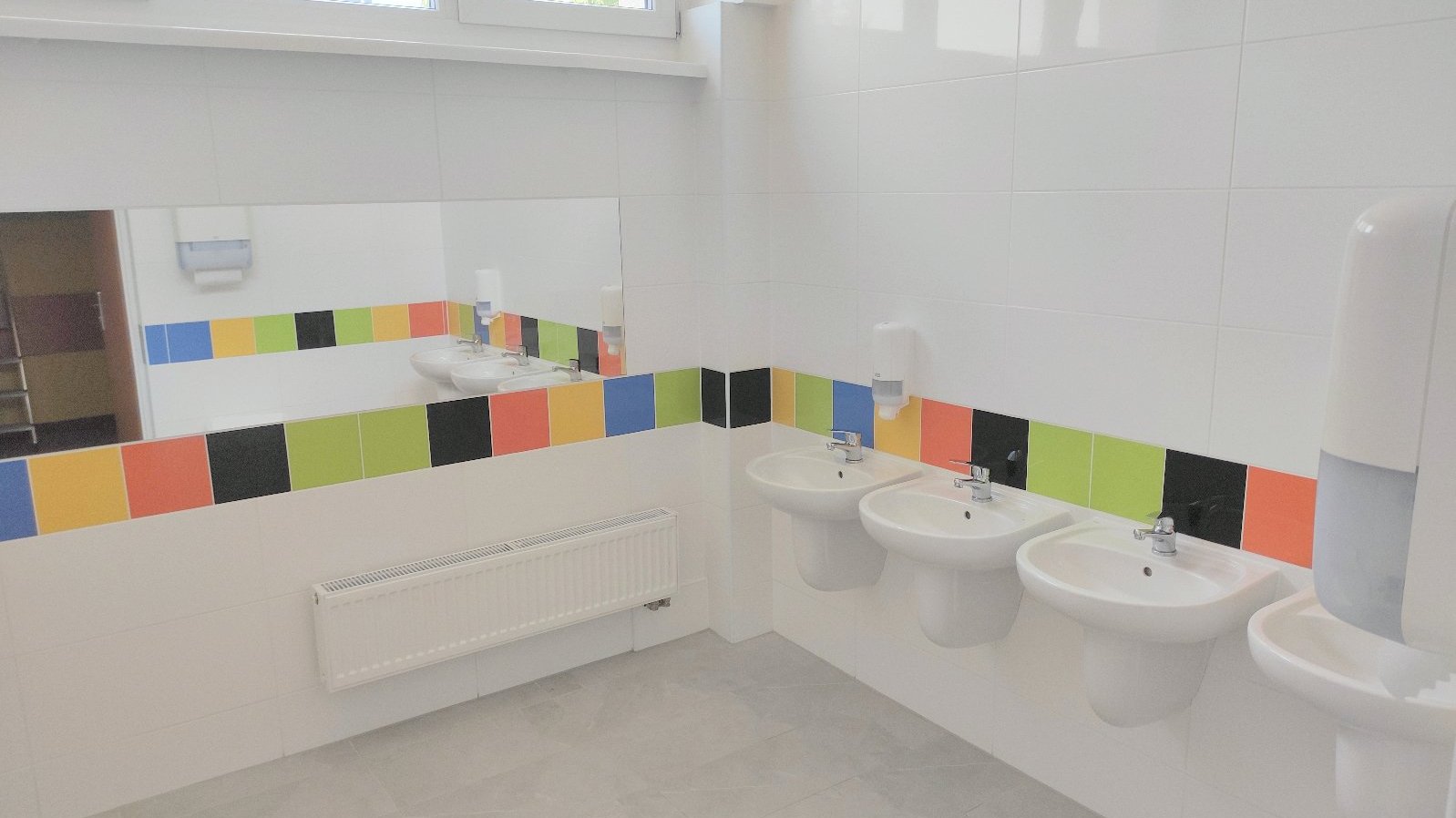 Zdjęcie przedstawia wyremontowaną łazienkę. Widać na nim stojące w rzędzie umywalki i lustro.