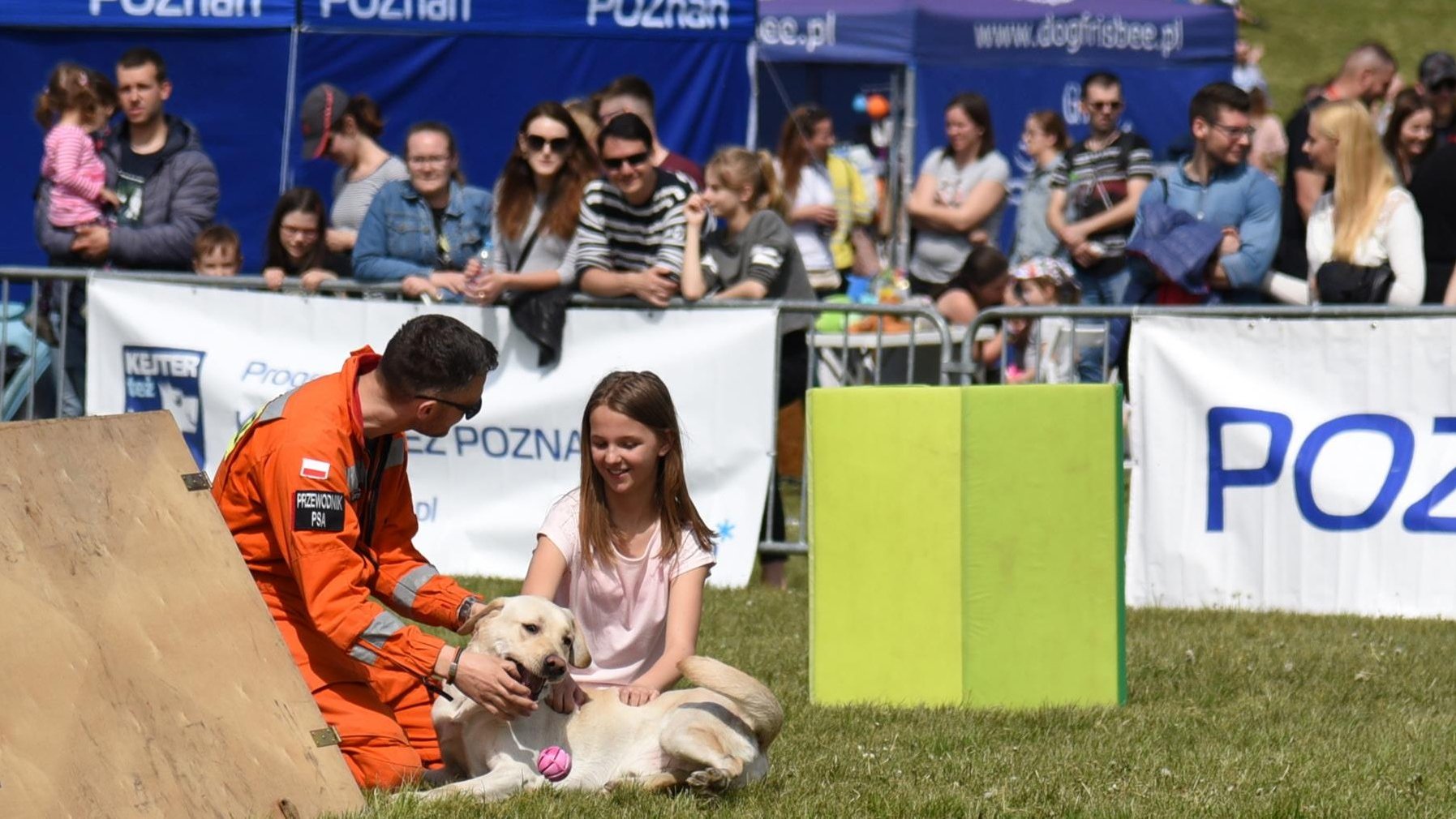 Zdjęcie przedstawia mężczyznę w pomarańczowym stroju z napisem "przewodnik psa", który klęczy na trawie w towarzystwie psa i małej dziewczynki. W tle, za barierką widać innych ludzi.
