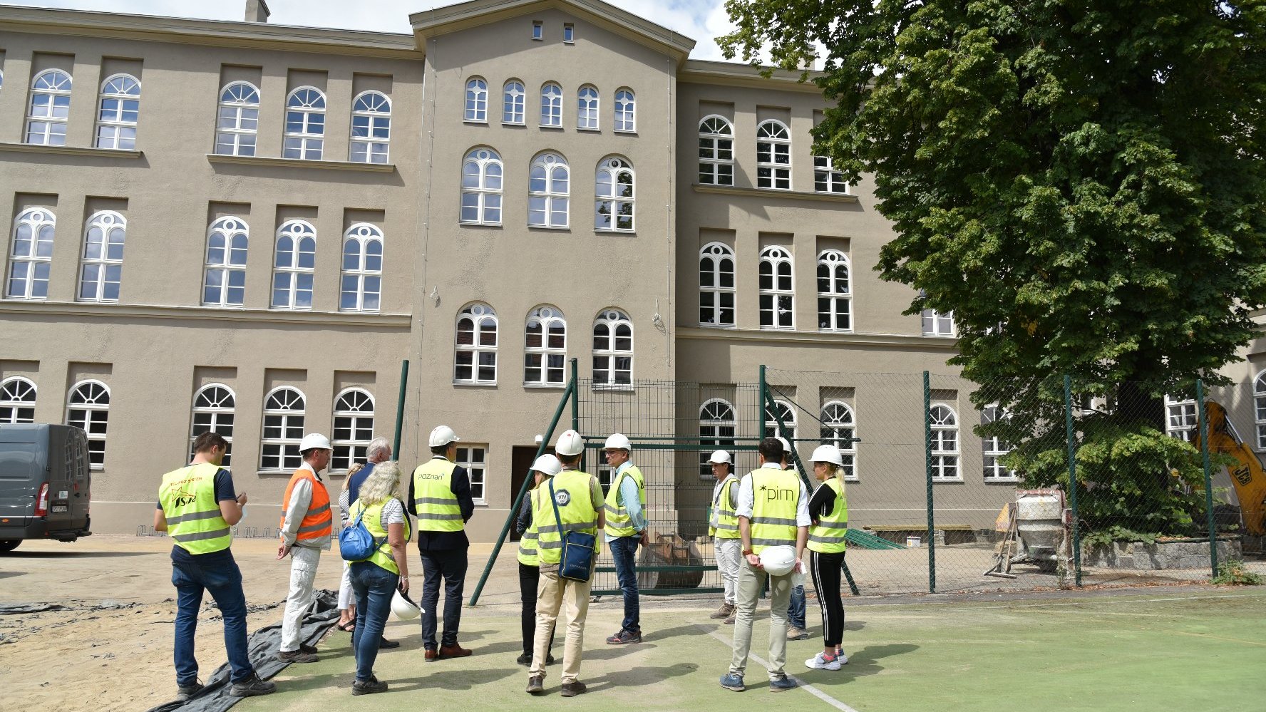 Zdjęcie przedstawia grupę ludzi w żółtych kamizelkach stojących przed remontowanym budynkiem szkoły. - grafika artykułu