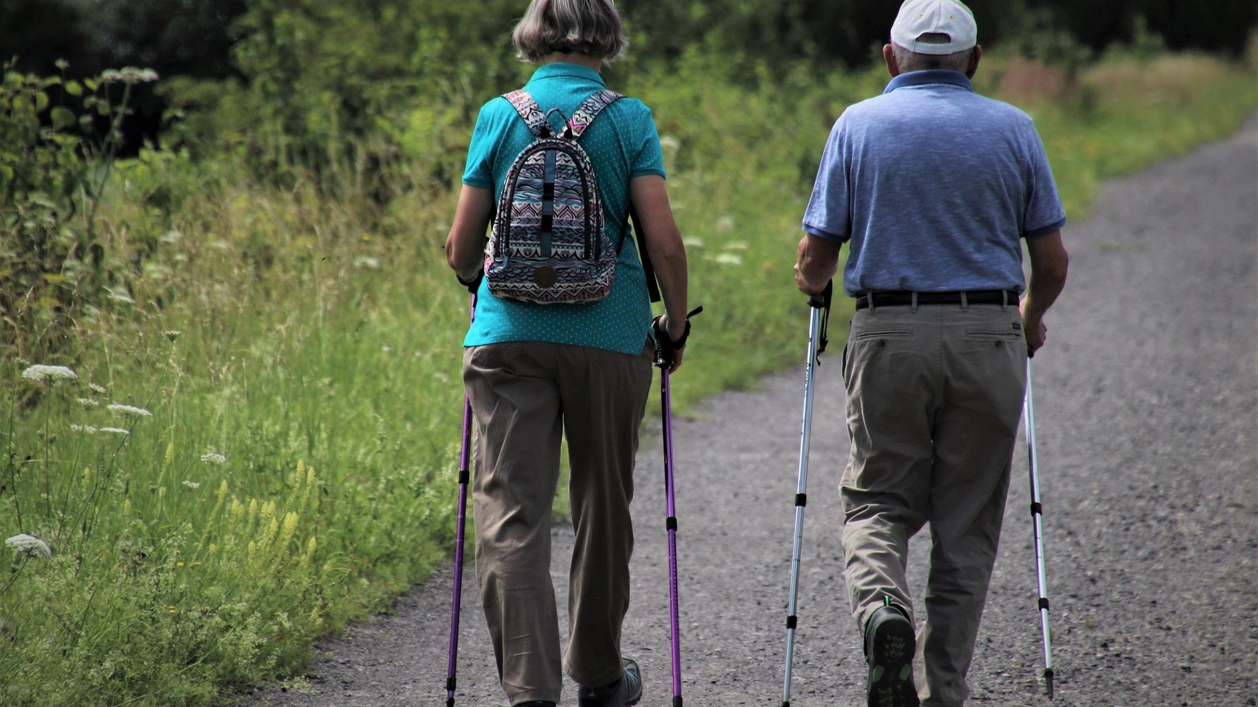 Na zdjęciu dwoje starszych ludzi, tyłem do obiektywu, maszerują z kijkami do nordic walking