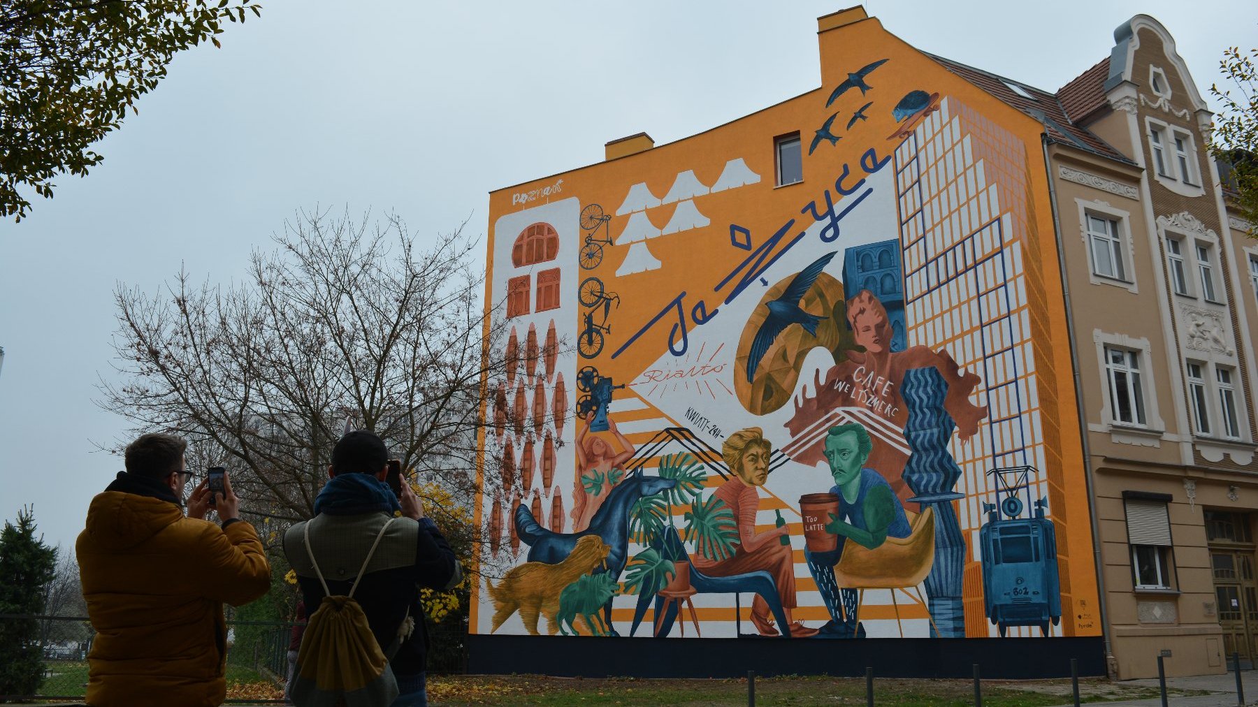 Galeria zdjęć przedstawia nowy mural na Jeżycach. Widać na nim m.in. dwóch poetów Słowackiego i Mickiewicza. Obok znajduje się ulica z samochodami.