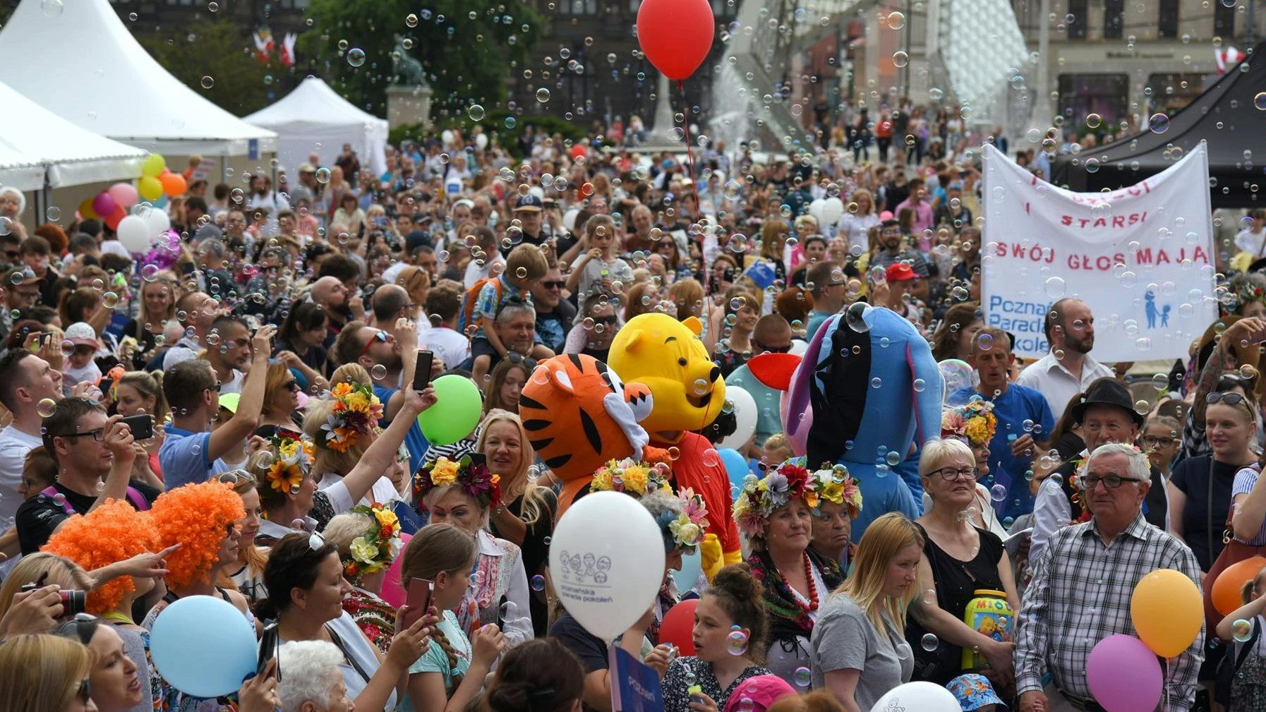 Na zdjęciu tłum uśmiechniętych ludzi, starszych, dorosłych i dzieci, niektórzy trzymają balony; nad nimi bańki mydlane