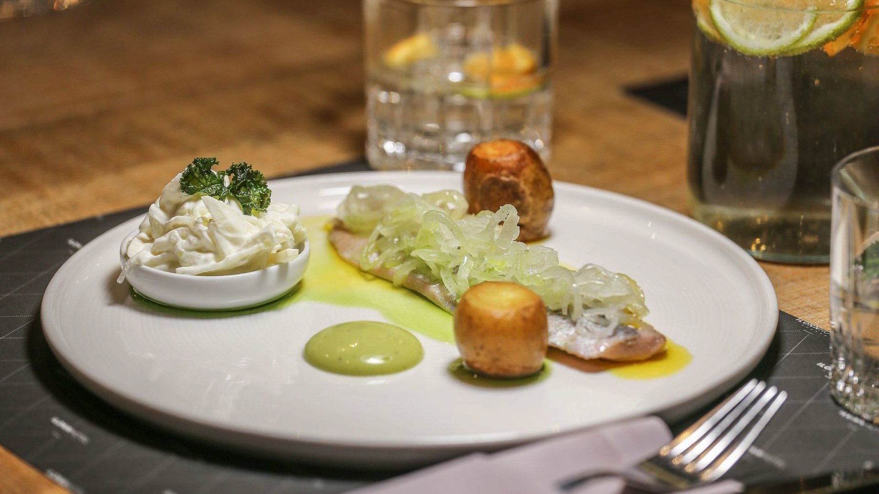 Na zdjęciu danie: kawałki ziemniaka, ryba i pojemnik z sosem, wszystko na białym talerzu. W tle szklanki i sztućce