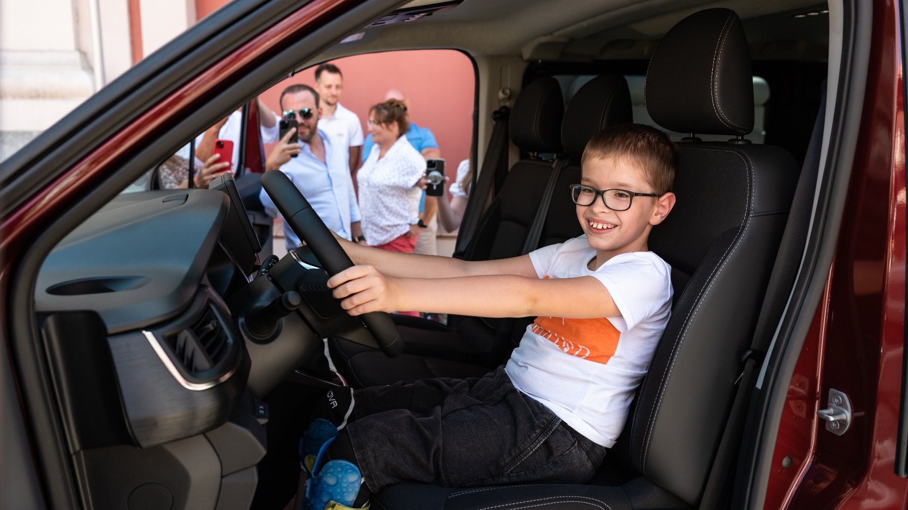 Na zdjęciu uśmiechnięty chłopiec w okularach siedzący za kierownicą samochodu