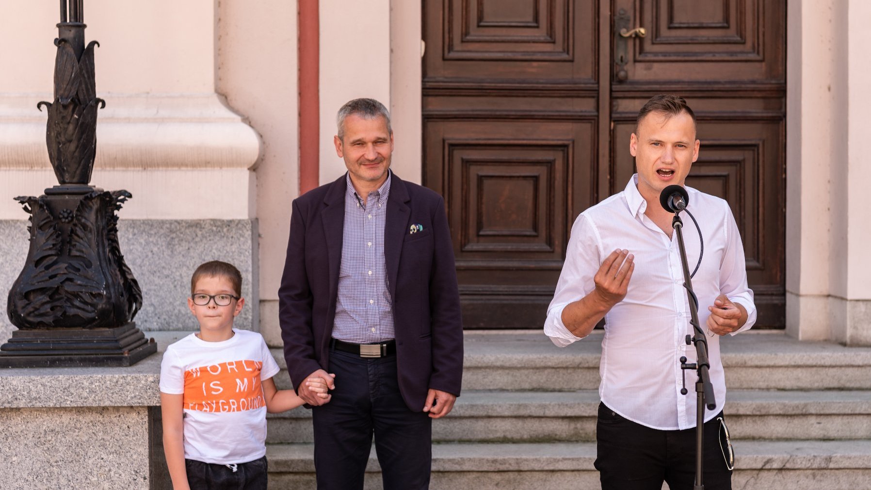 Na zdjęciu mężczyzna w białej koszuli mówi przez telefon, w tle stoi zastępca prezydenta Poznania i mały chłopiec