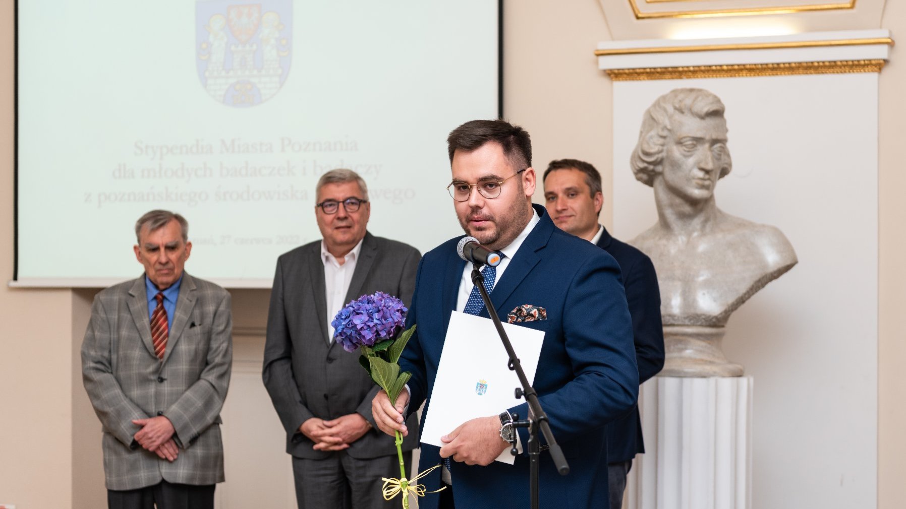 Na zdjęciu stypendysta przy mikrofonie, za nim zastępca prezydenta Poznania, przewodniczący rady miasta i przewodniczący kapituły