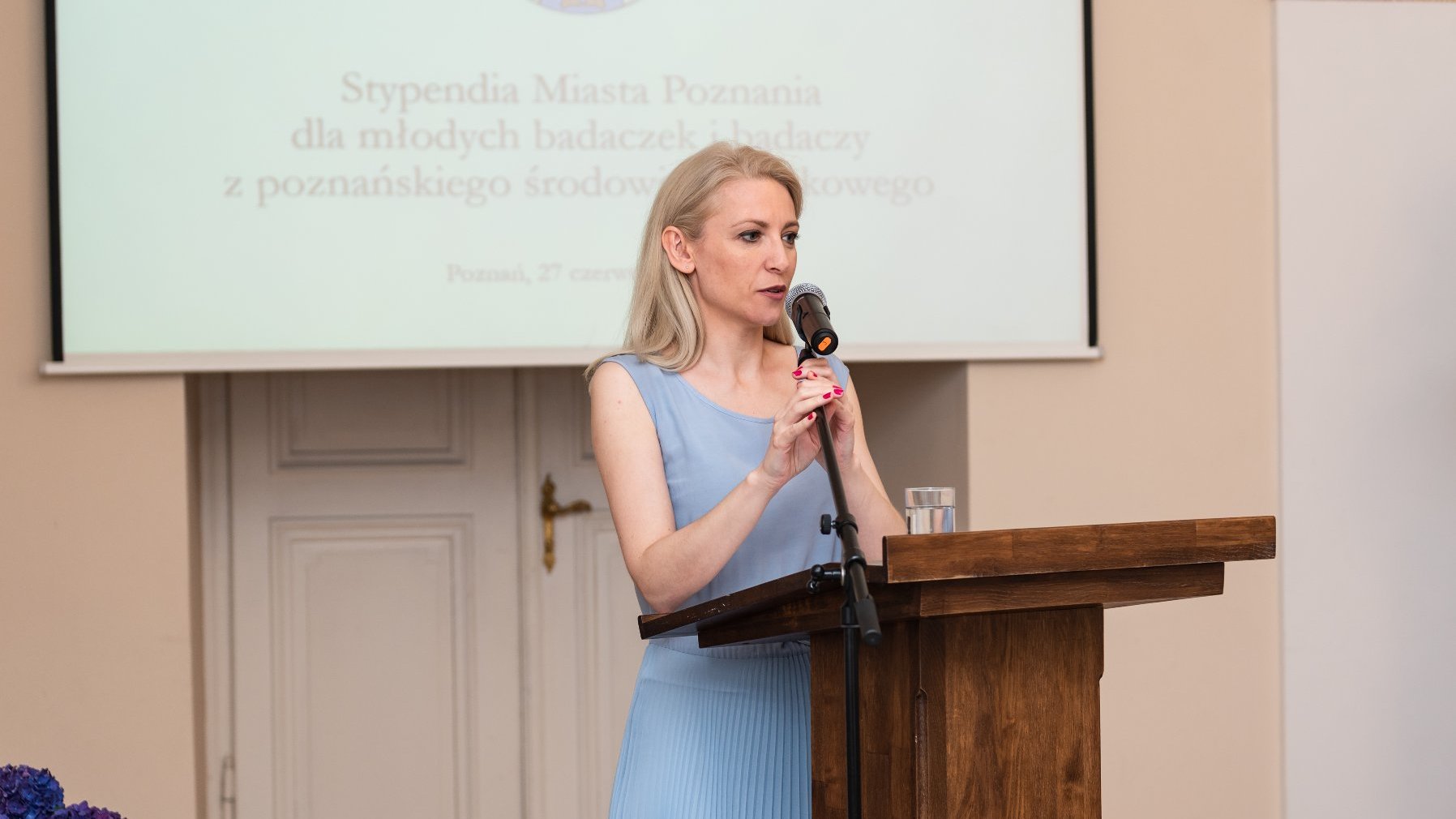 Na zdjęciu kobieta w błękitnej sukni, stoi przy mównicy z mikrofonem w ręku