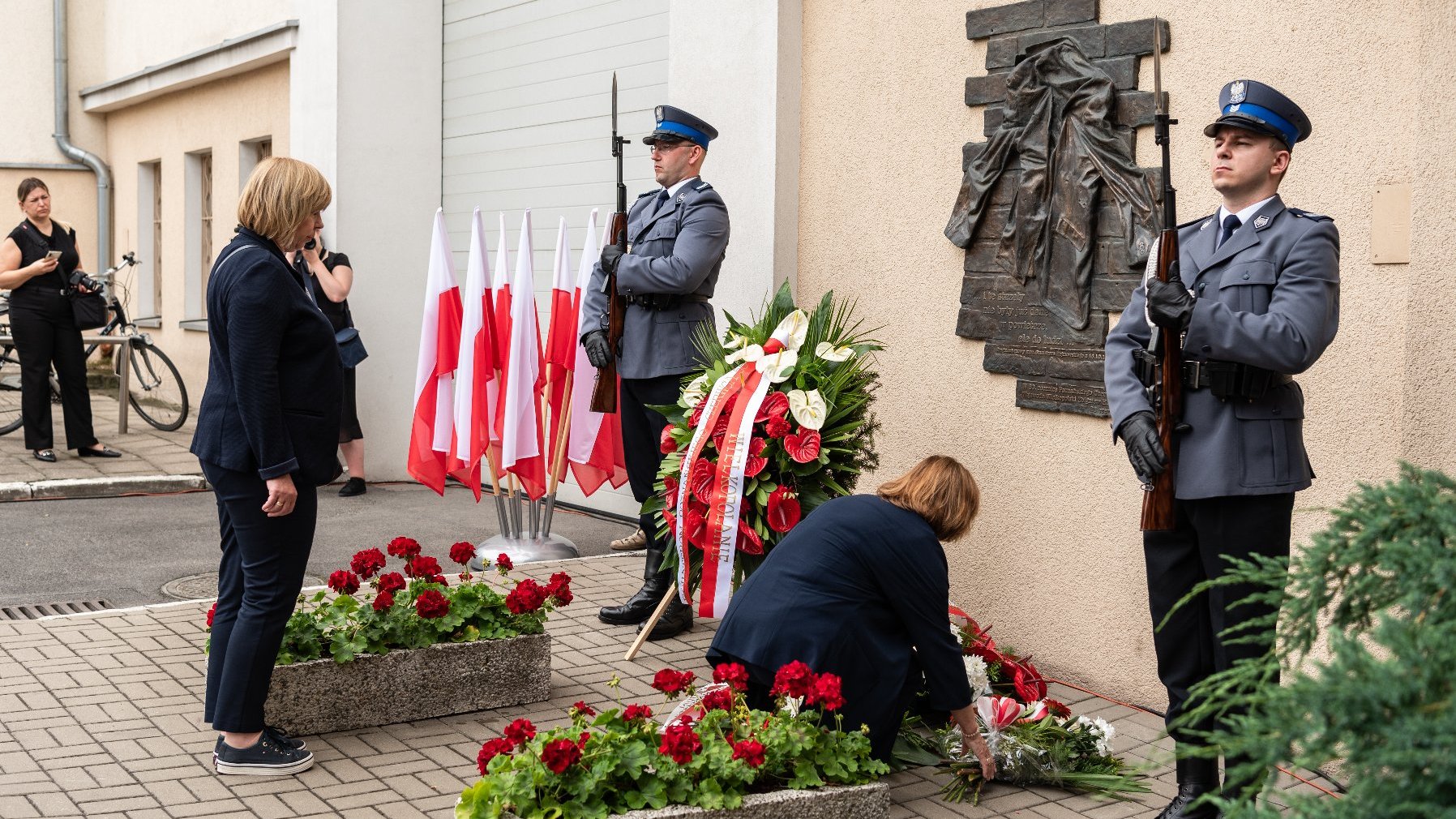 Na zdjęciu składanie kwiatów pod komendą policji, przy tablicy asystują dwaj policjanci