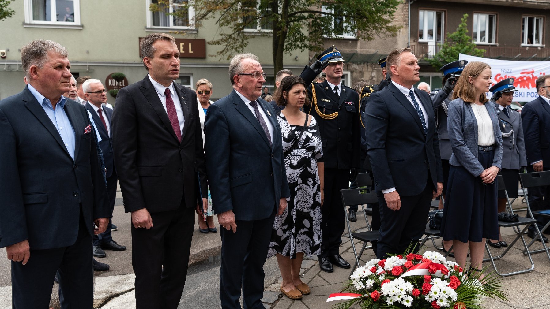 Na zdjęciu uczestnicy uroczystości stojący obok siebie i śpiewający hymn, mężczyźni w mundurach salutują