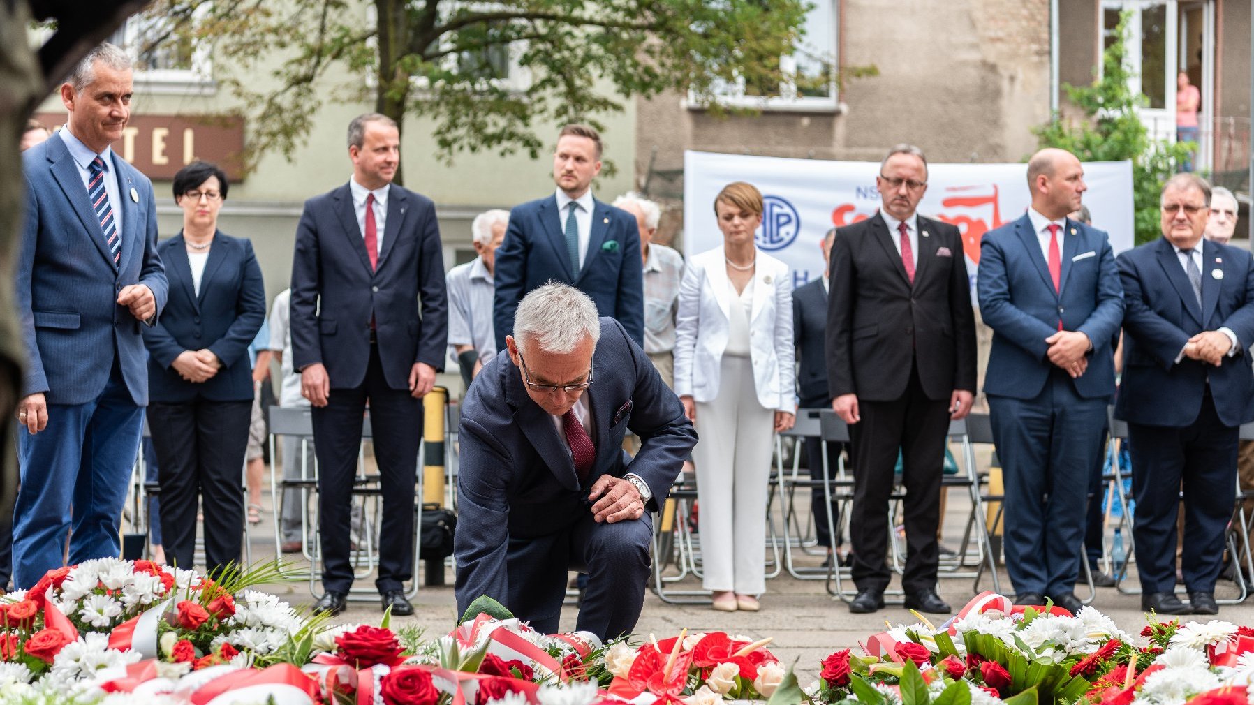 Na zdjęciu mężczyzna, przyklęka i składa kwiaty przed tablicą, w tle widać pozostałych uczestników wydarzenia