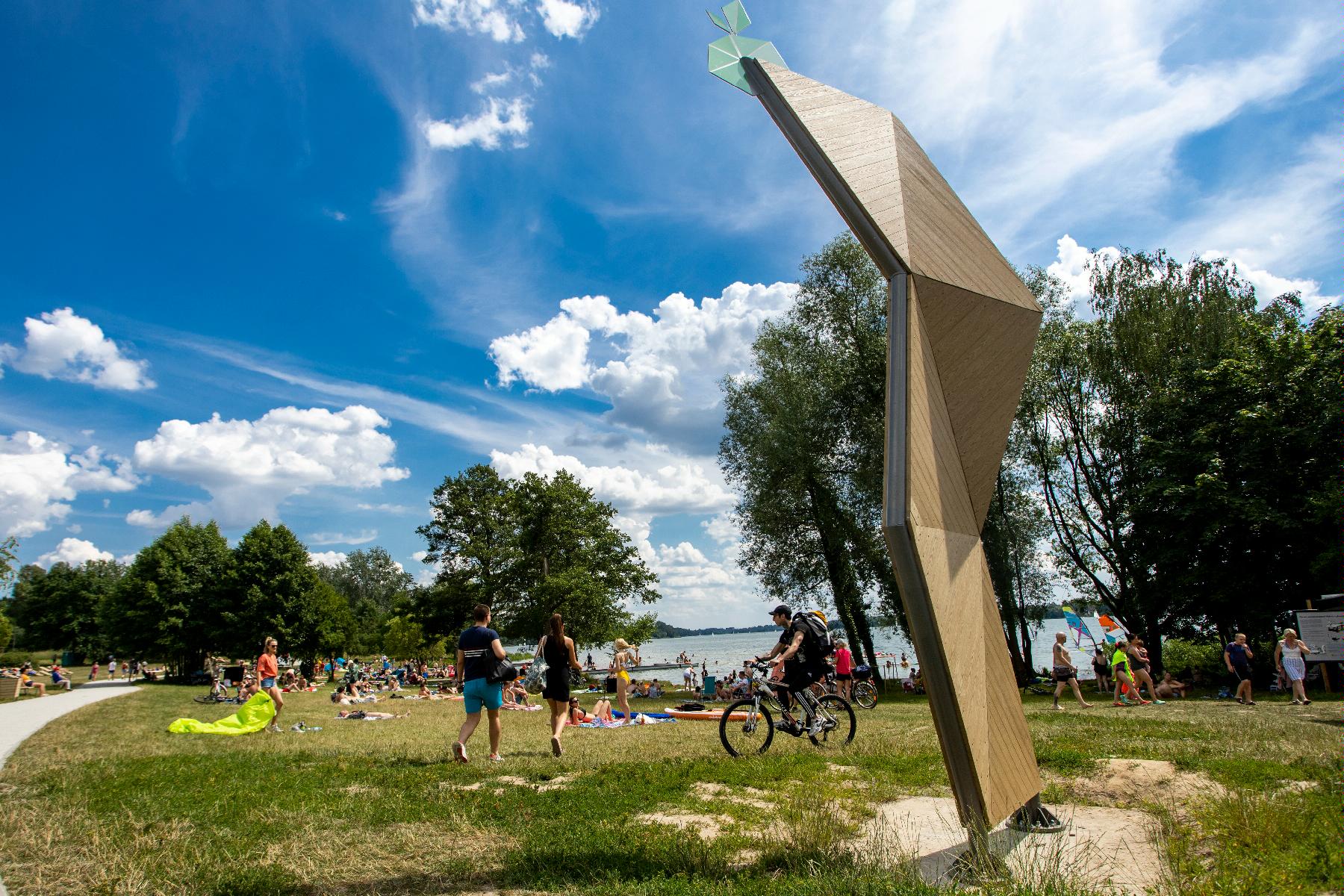 Zdjęcie przedstawia plażę nad jeziorem Kierskim. Widać na nim ludzi opalających się, spacerujących i jeżdżących na rowerach. W tle znajduje się woda. - grafika artykułu