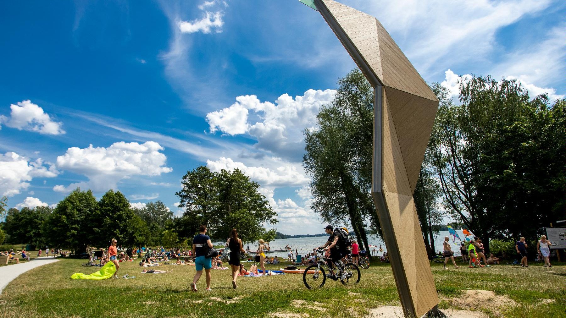 Zdjęcie przedstawia plażę nad jeziorem Kierskim. Widać na nim ludzi opalających się, spacerujących i jeżdżących na rowerach. W tle znajduje się woda.