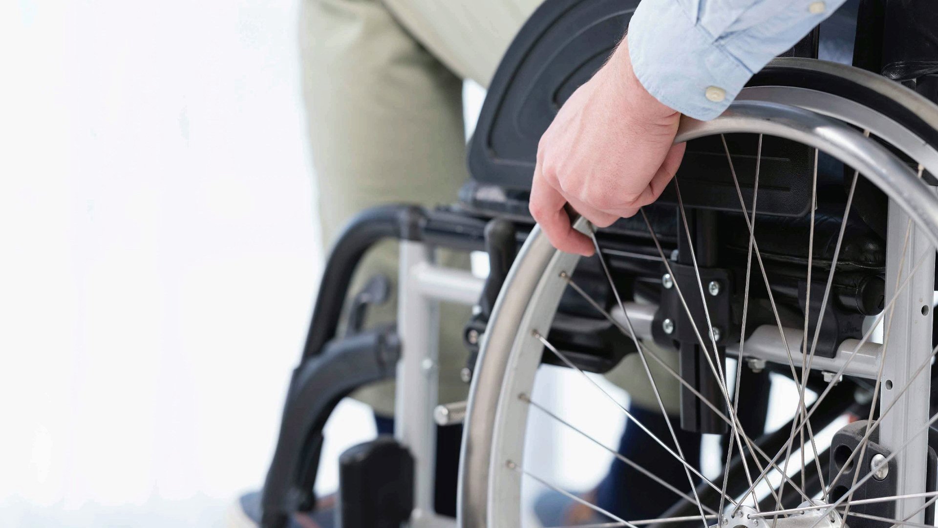 Na zdjęciu mężczyzna siedzący na wózku inwalidzkim, widać tylko dolną część ciała, w centrum dłoń na kole wózka