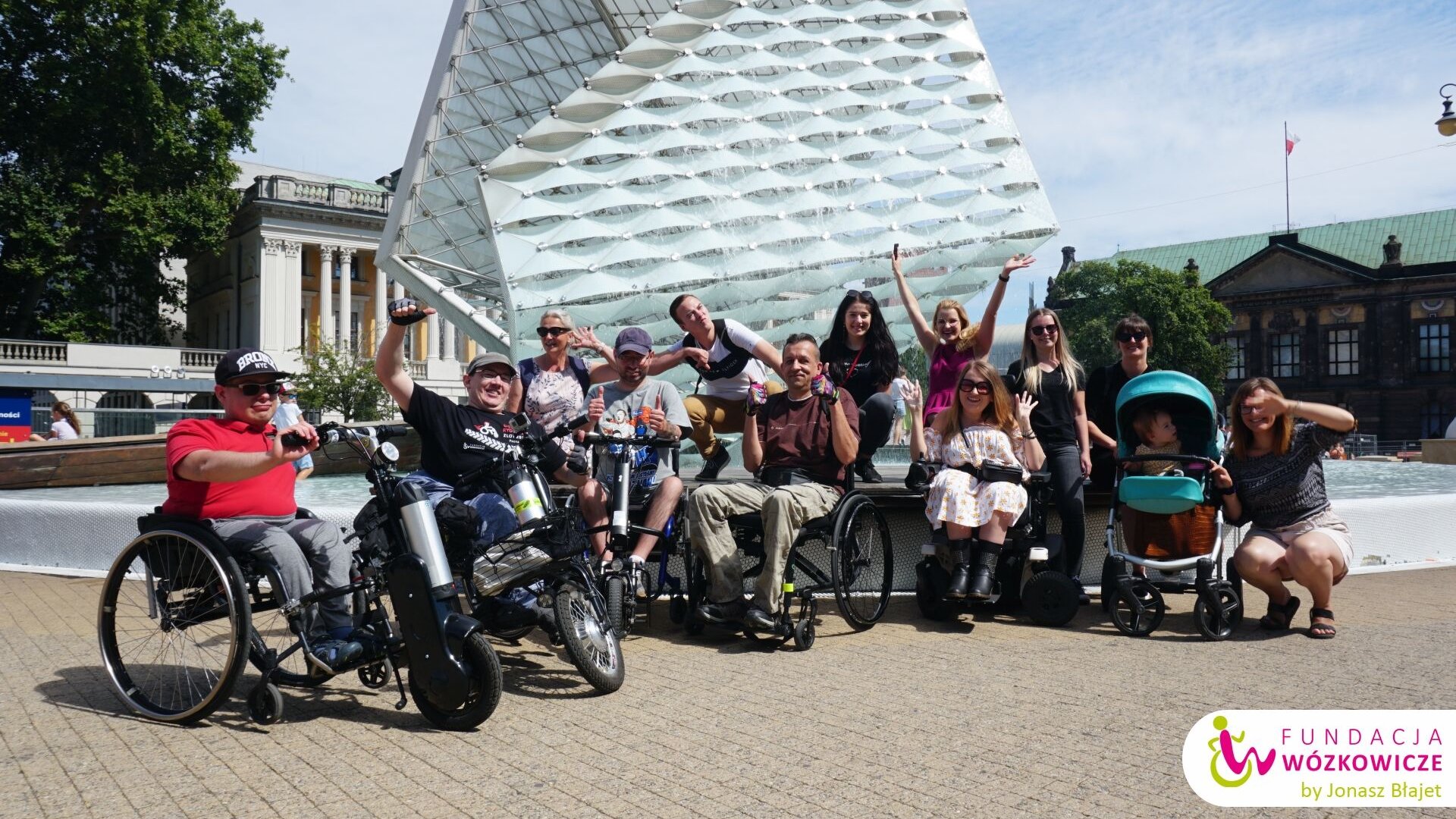 Zdjęcie przedstawia grupę osób z niepełnosprawnościami na wózkach, w tle fontanna na pl. Wolności.