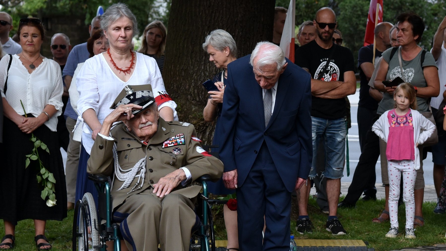 Na zdjęciu dóch starszych mężczyzn, jeden w mundurze i na wózku, salutuje, drugi w garniturze