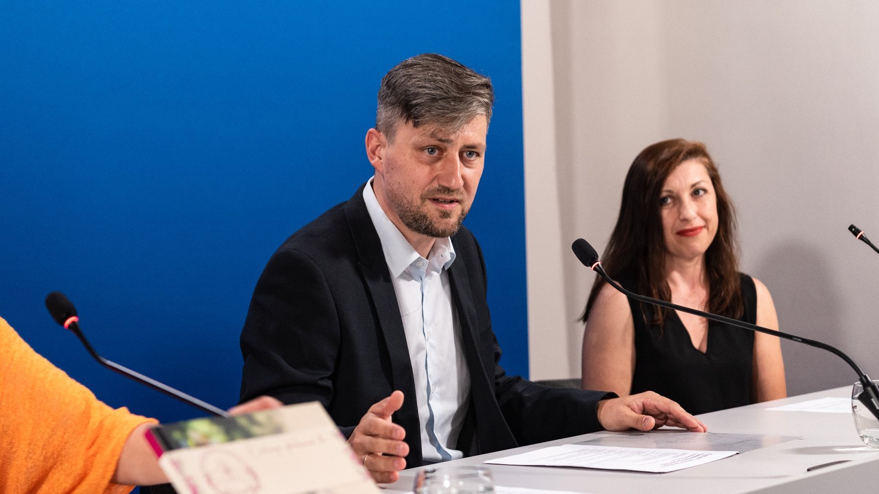 Na zdjęciu dwie osoby - mężczyzna i kobieta - za stołem konferencyjnym, przy mikrofonach