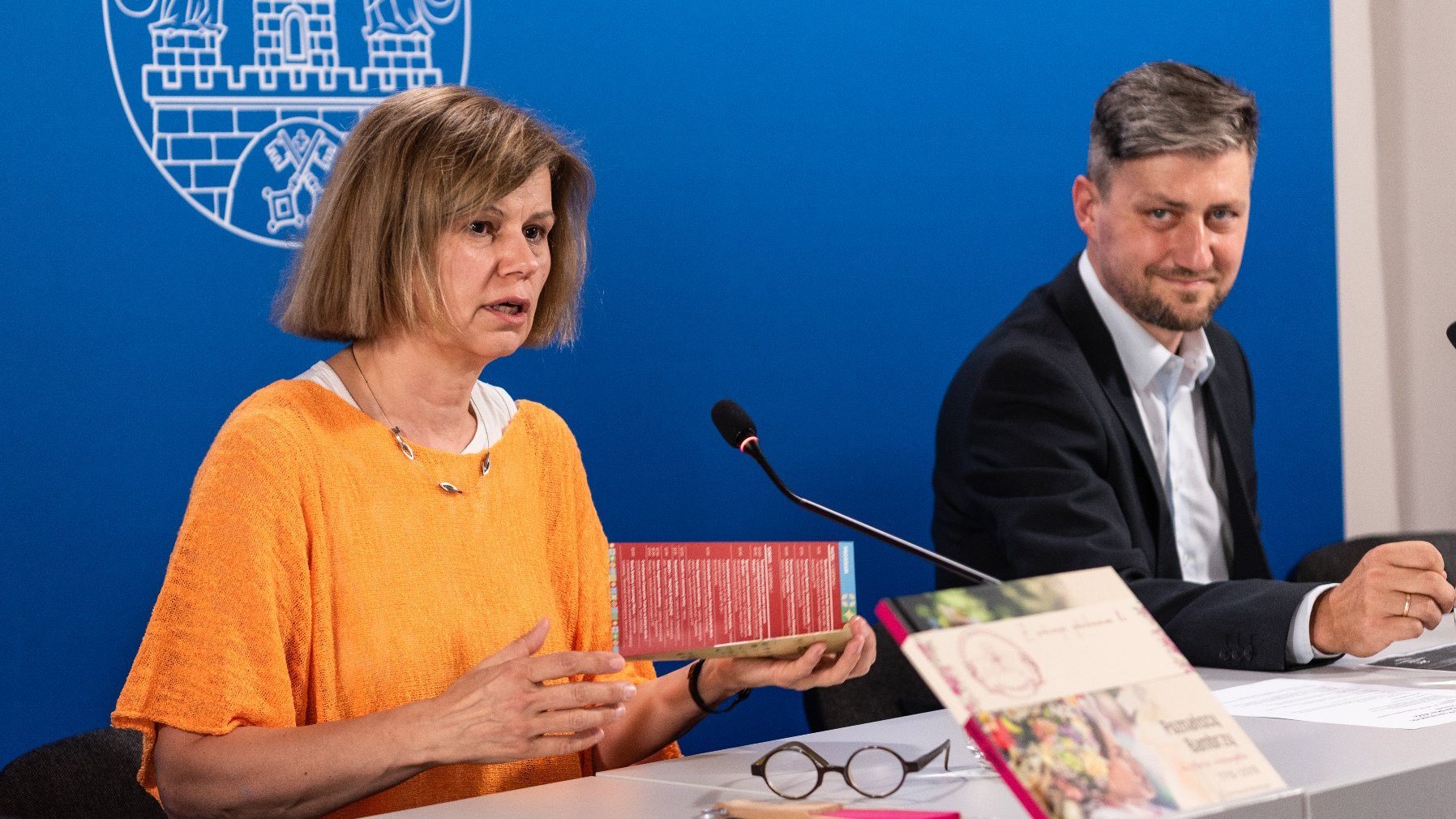 Na zdjęciu dwie osoby za stołem konferencyjnym, zwrócone do obiektywu półprofilem, mężczyzna i kobieta