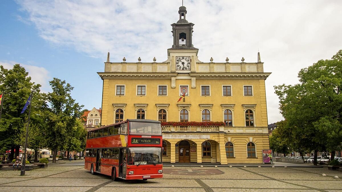 Na zdjęciu czerwony piętrowy autobus, stojący przed renesansowym ratuszem