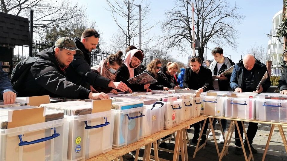 Na zdjęciu grupa ludzi przeglądająca wystawione na sprzedaż winyle, w tle widać drzewa