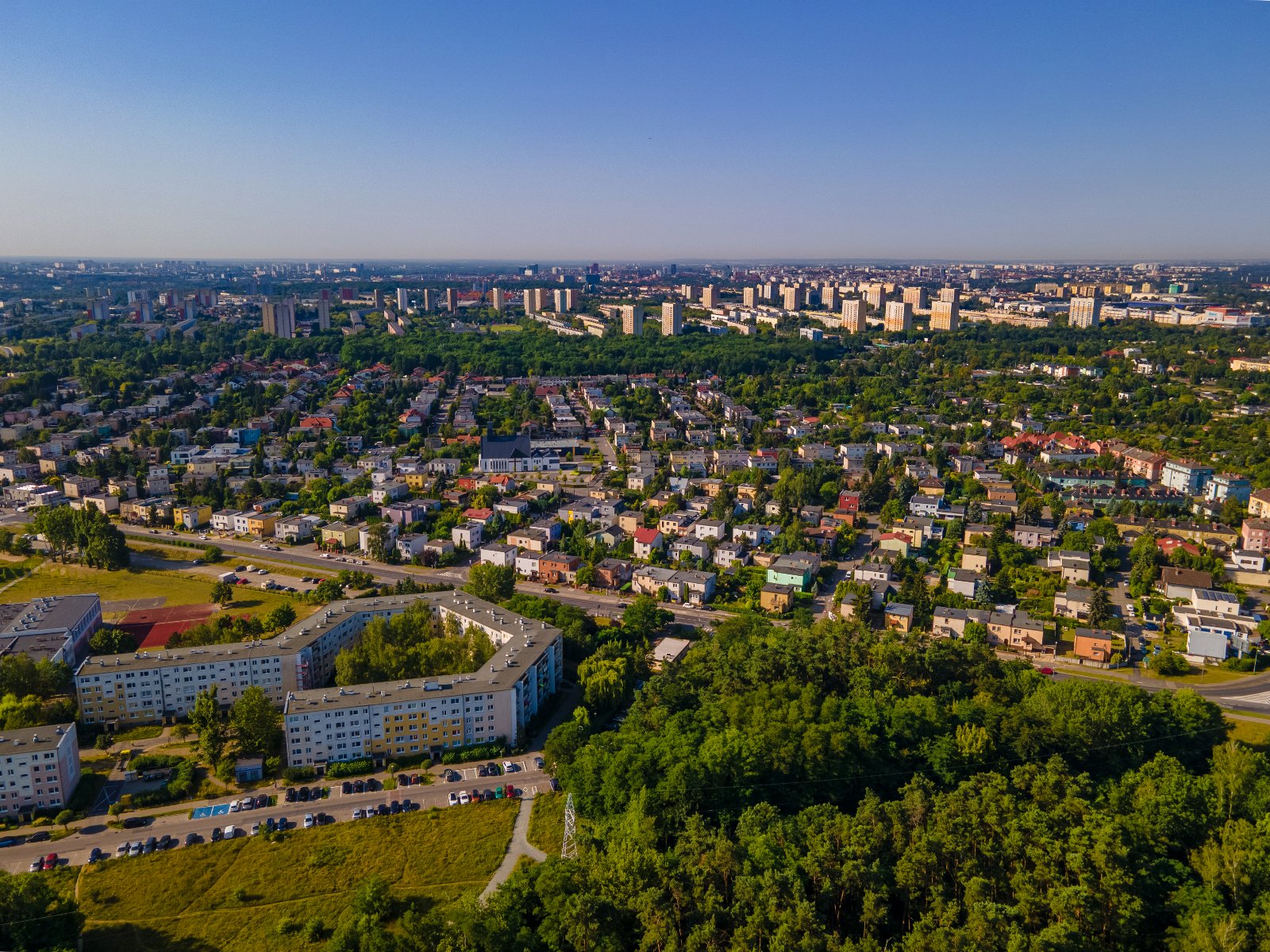 Zdjęcie z lotu ptaka przedstawia zieleń (na dole), obok bloki, później pas domków jednorodzinnych, a dalej bloki mieszkalne - grafika artykułu