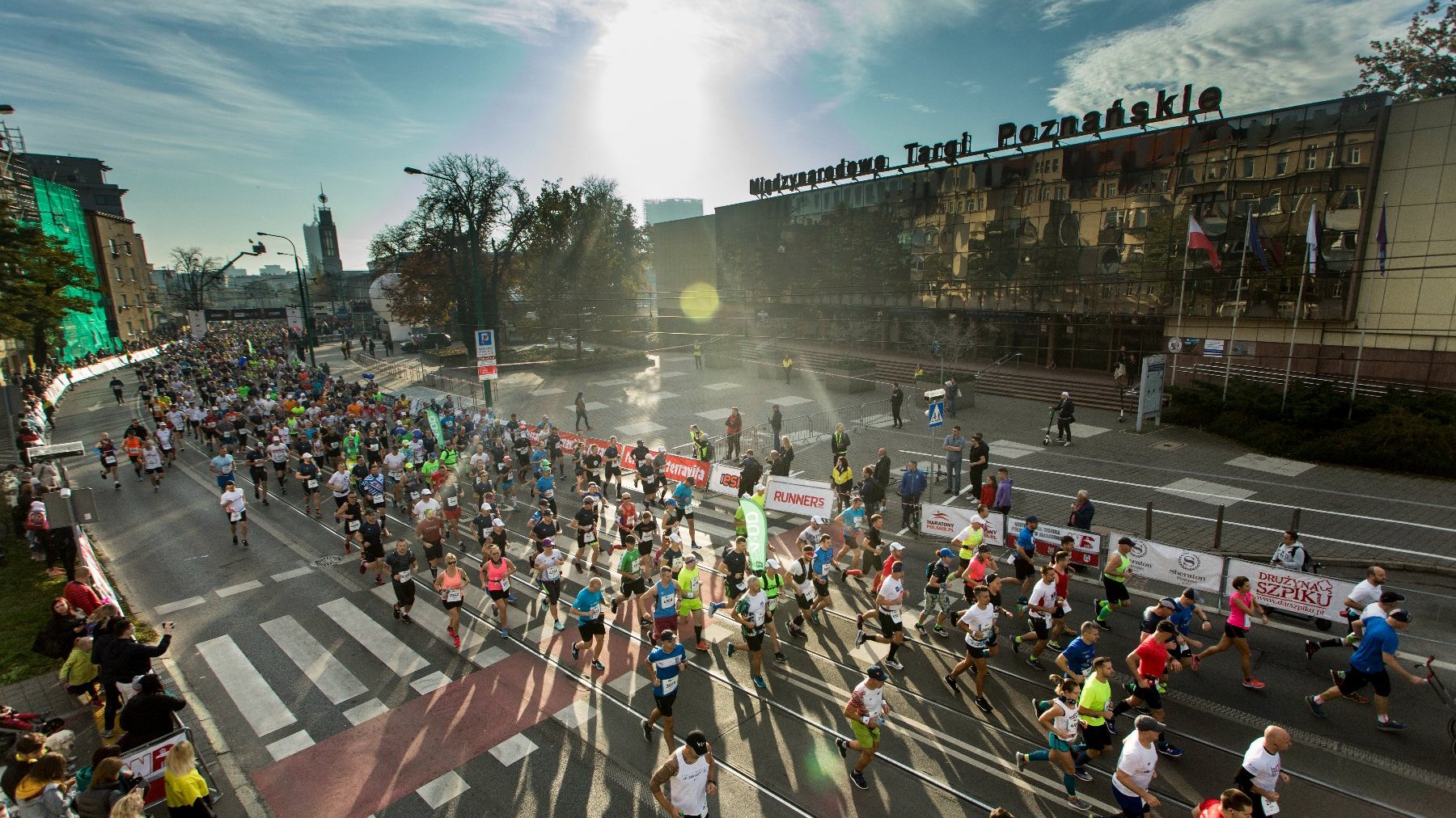 Na zdjęciu ludzie biegnący ulicami Poznania podczas maratonu, widok z lotu ptaka; w tle widać budynek MTP.