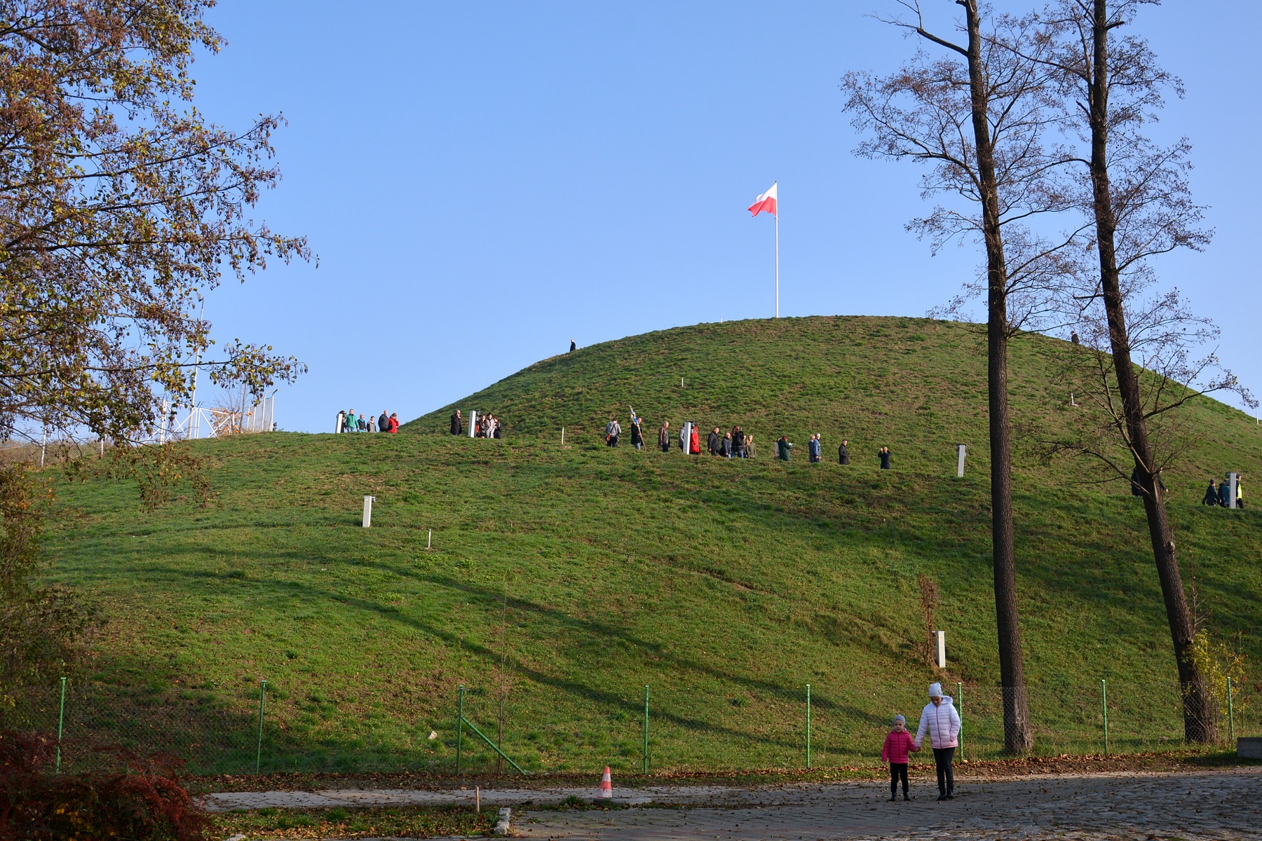 Zdjęcie przedstawia Kopiec Wolności w Poznaniu oraz wchodzących na niego ludzi. Na szczycie widąć flagę Polski. - grafika artykułu