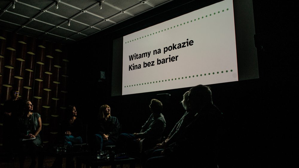 Grupa osób w sali kinowej patrzy na ekran, na któyrm wyświetlony jest napis "Witamy na pokazie Kina bez barier".