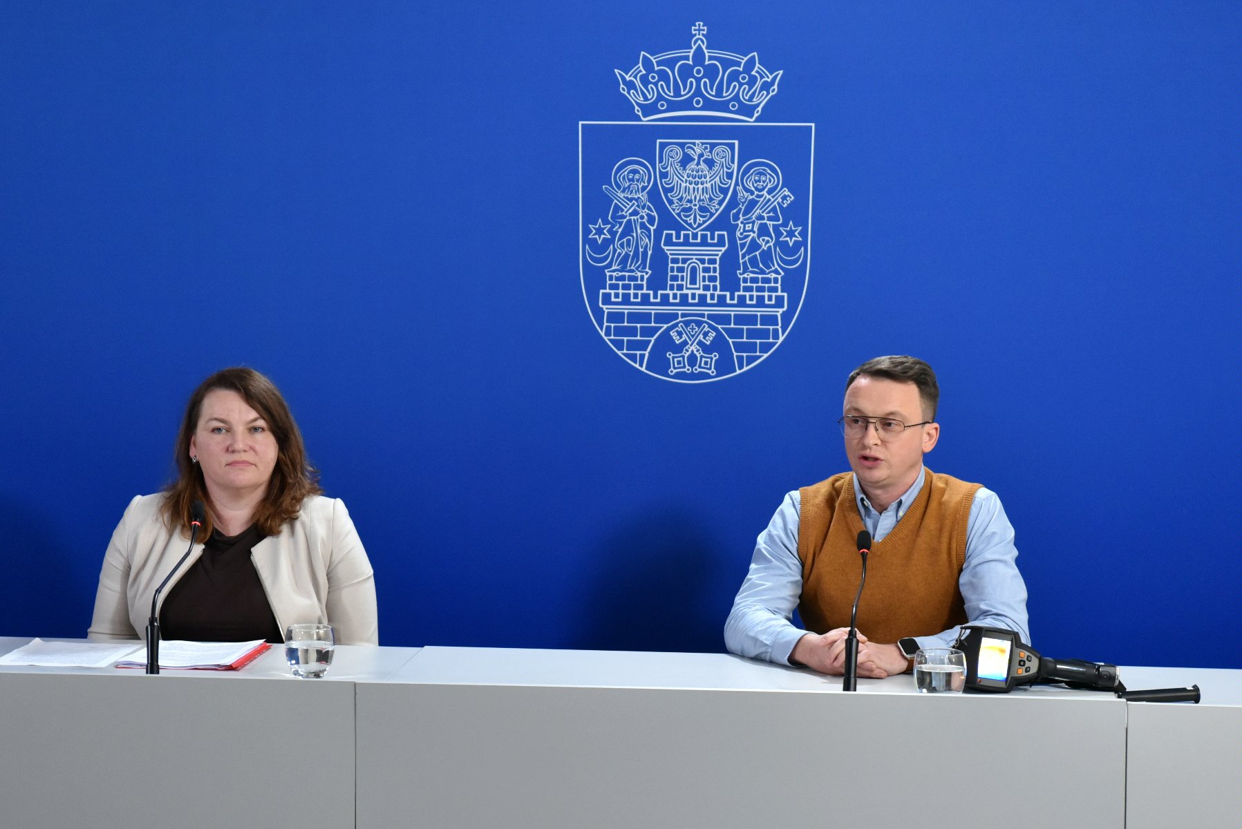 Dwie osoby za stołem konferencyjnym, mężczyzna i kobieta, za nimi granatowa ścianka z herbem miasta - grafika artykułu