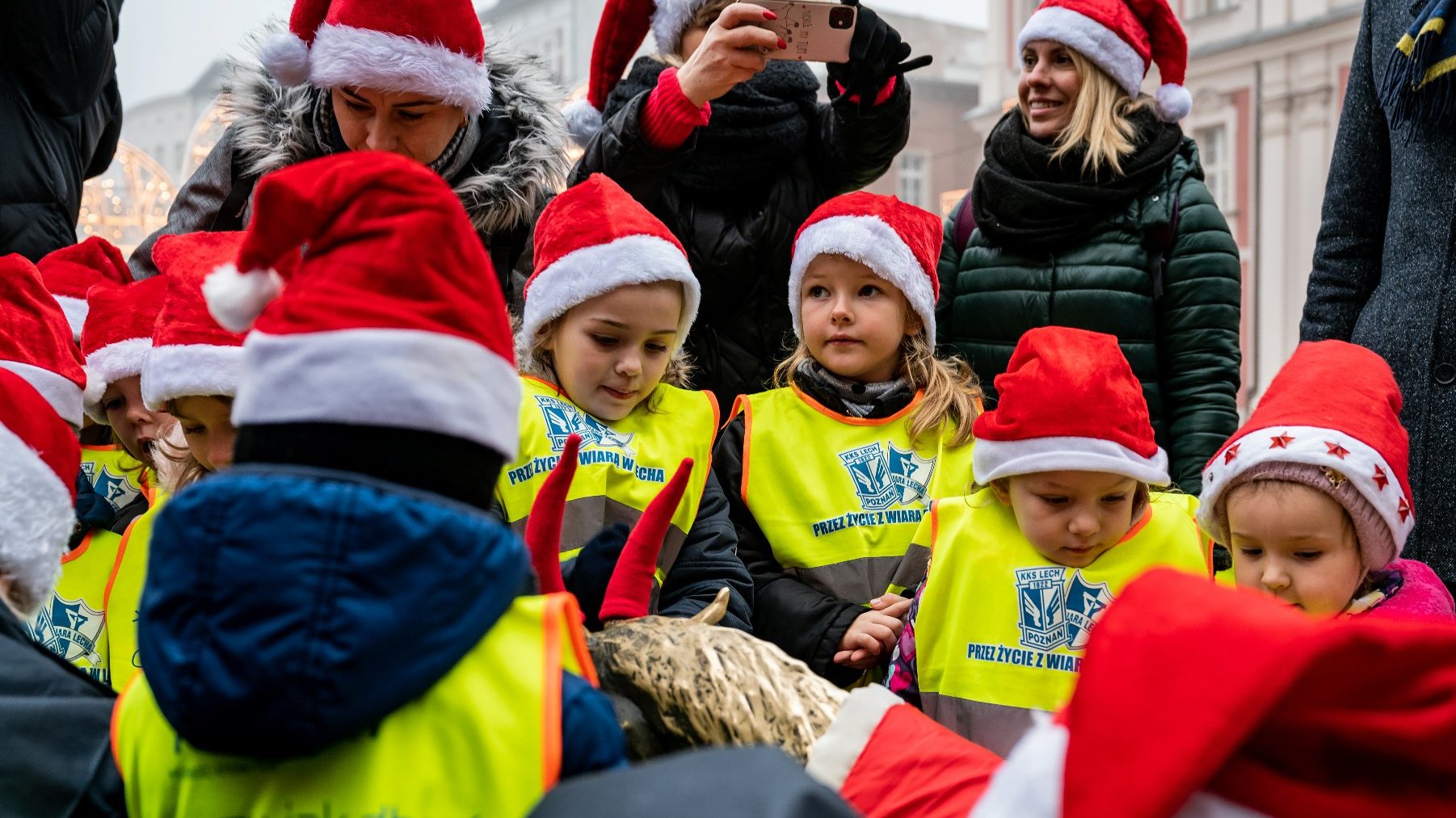 Galeria zdjęć przedstawia grupę dzieci i dorosłych dekorujących rzeźbę koziołków w mikołajkowe stroje.