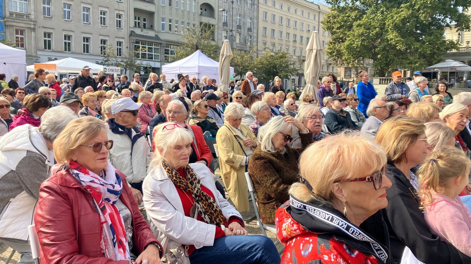 Na zdjęciu grupa seniorów w przestrzeni miasta, podczas jakiegoś miejskiego wydarzenia